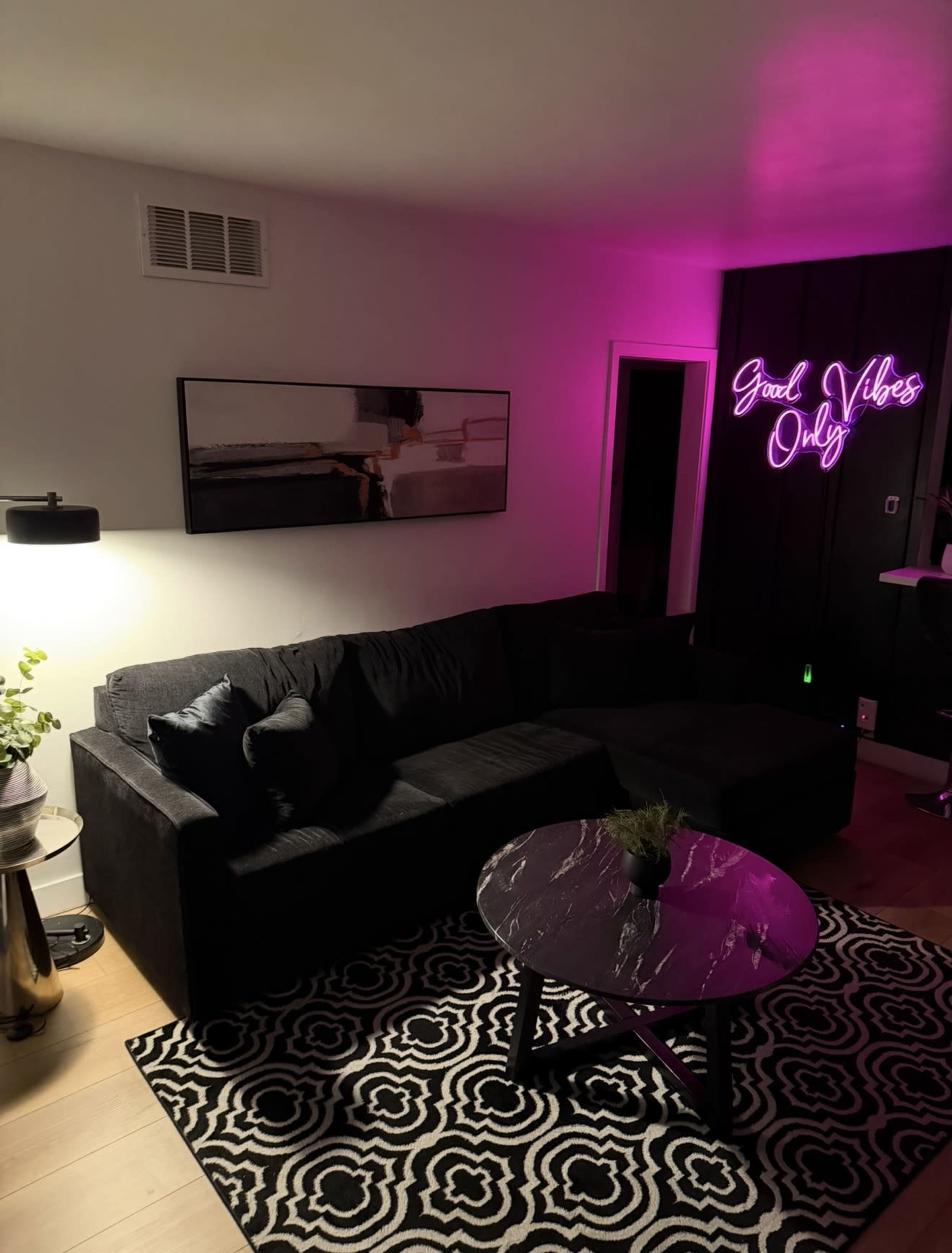 A modern living room features a black sectional sofa, a marble coffee table, and a neon sign that reads "Good Vibes Only," illuminated by purple light.