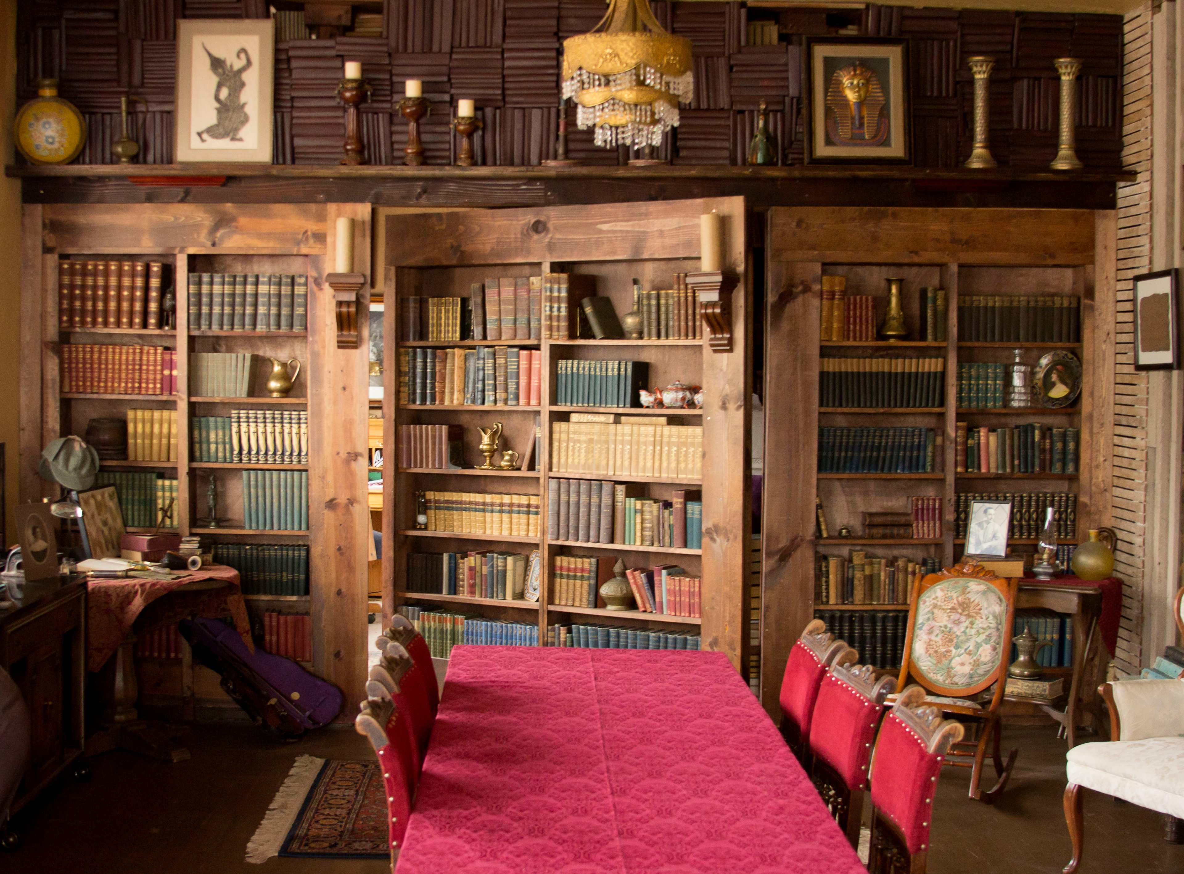 A room featuring a wooden bookshelf filled with books, framed art on the walls, and a large table with red chairs in front of it.