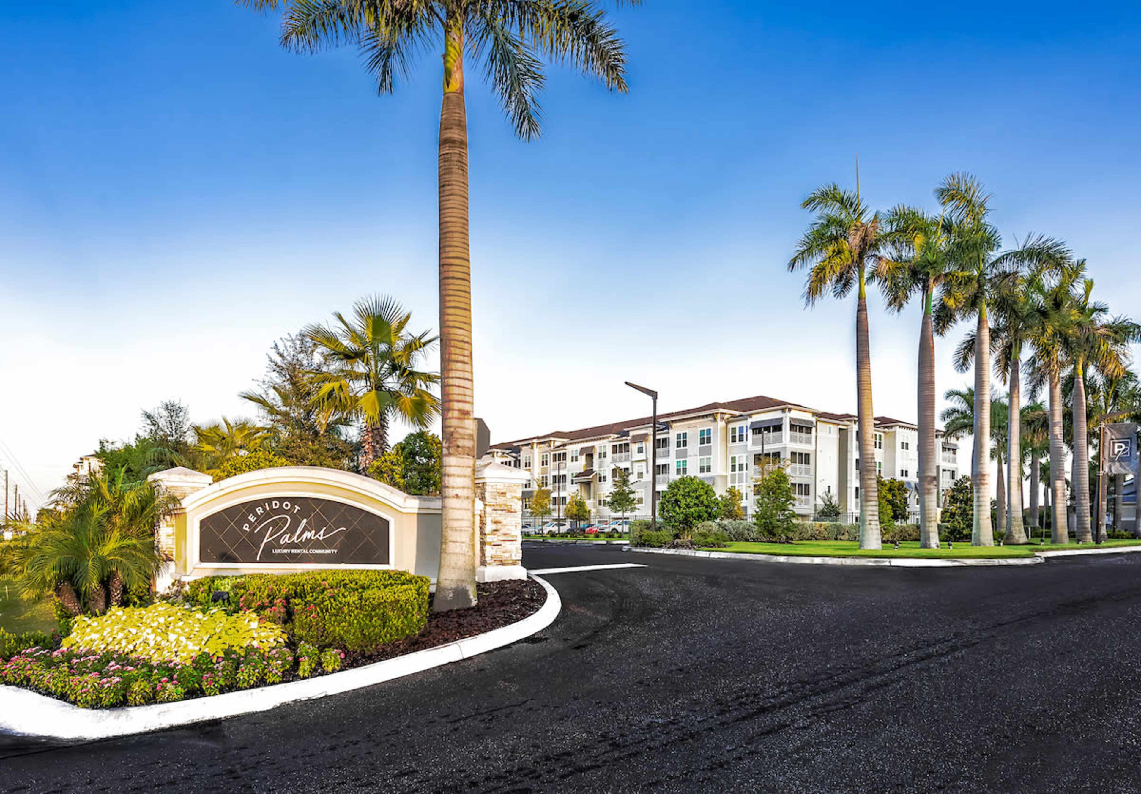 An entrance sign for a residential community surrounded by palm trees and landscaped gardens, with apartment buildings in the background.