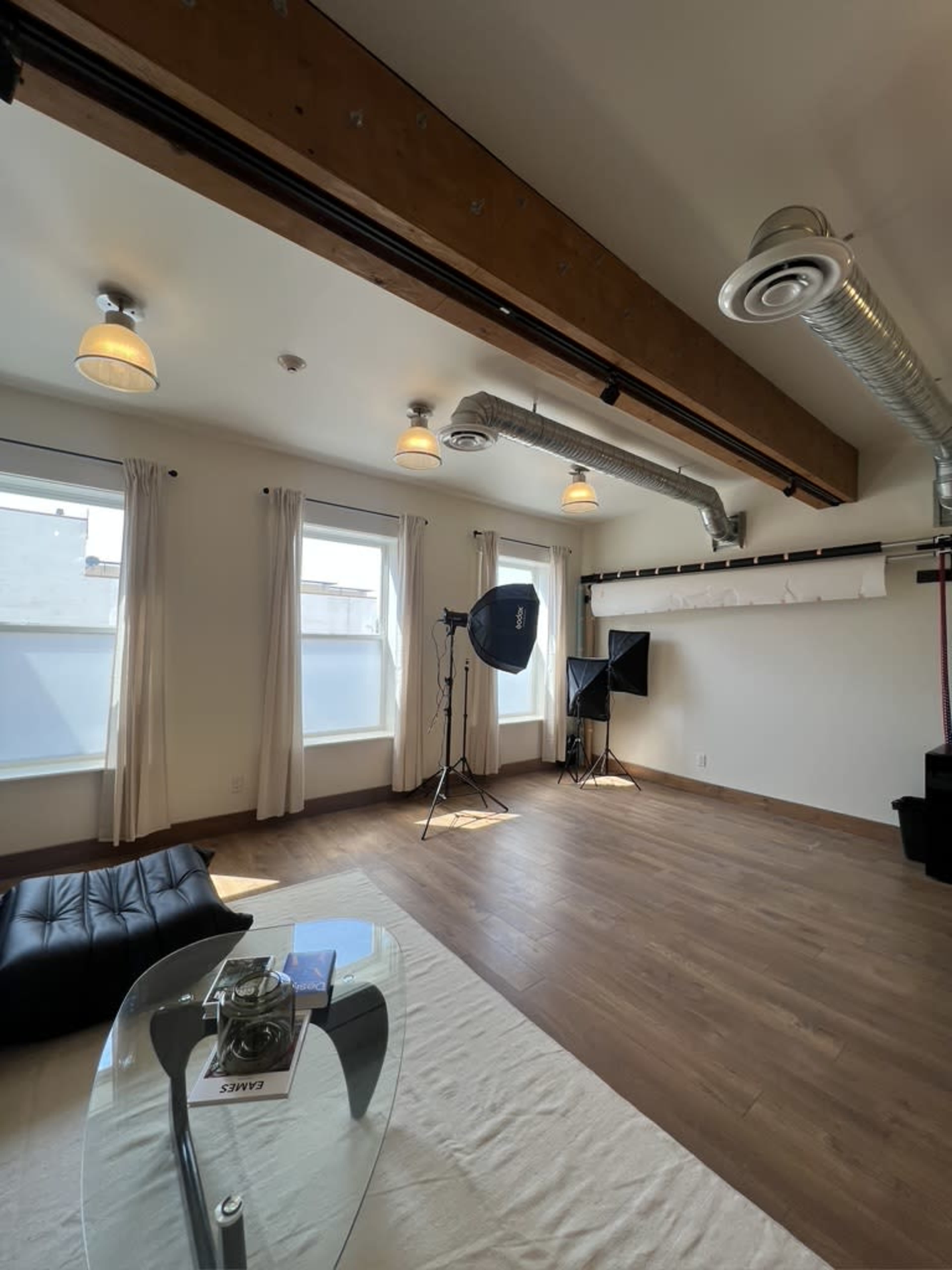 A spacious room with wooden floors, large windows, two light stands with softboxes, and minimal furnishings including a black couch and a glass table.
