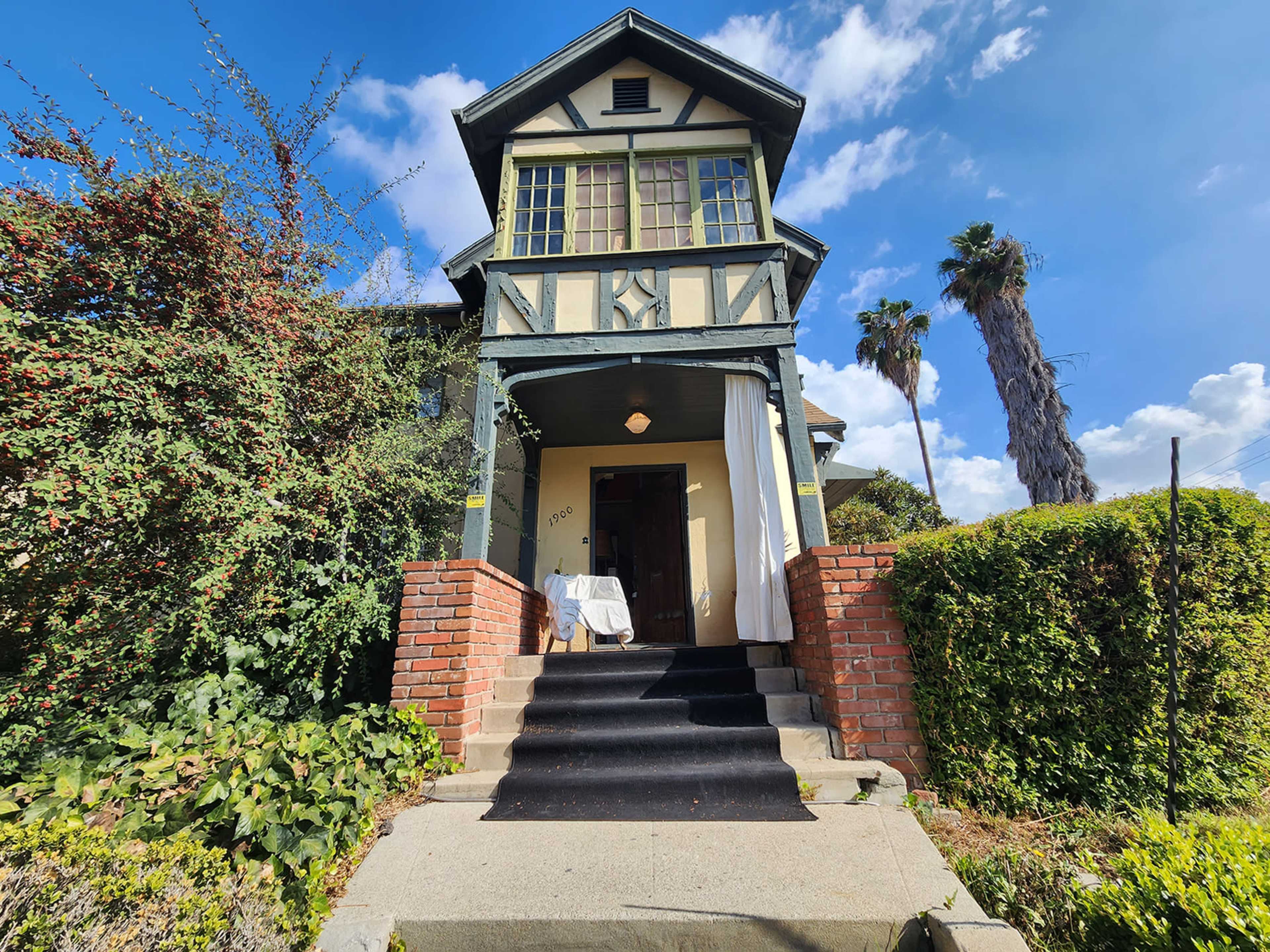 A two-story Tudor-style house with a prominent entrance and stairs leading up to a door, surrounded by greenery and palm trees.