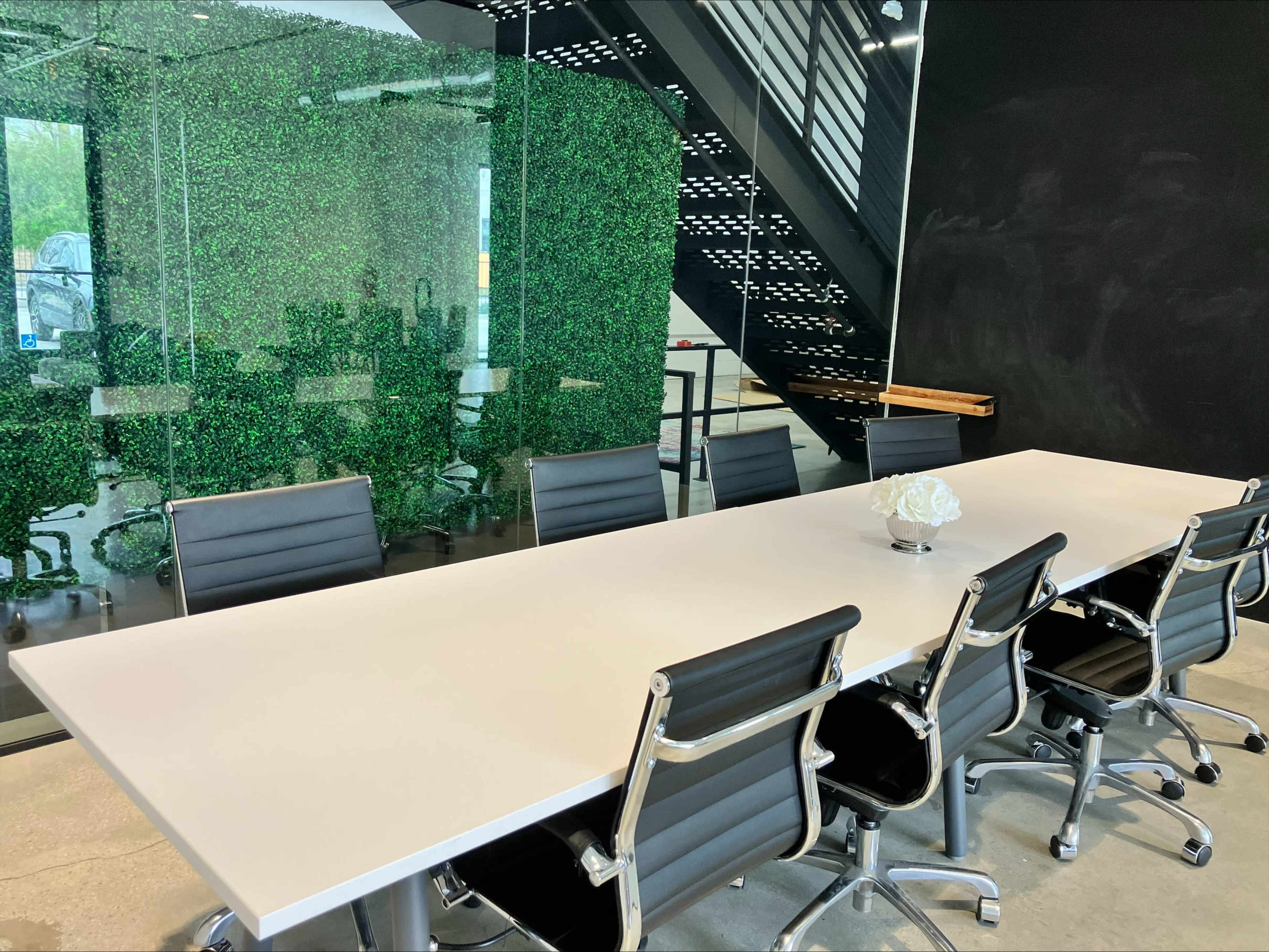 A modern conference room features a long white table surrounded by black chairs and a green wall of plants in the background.