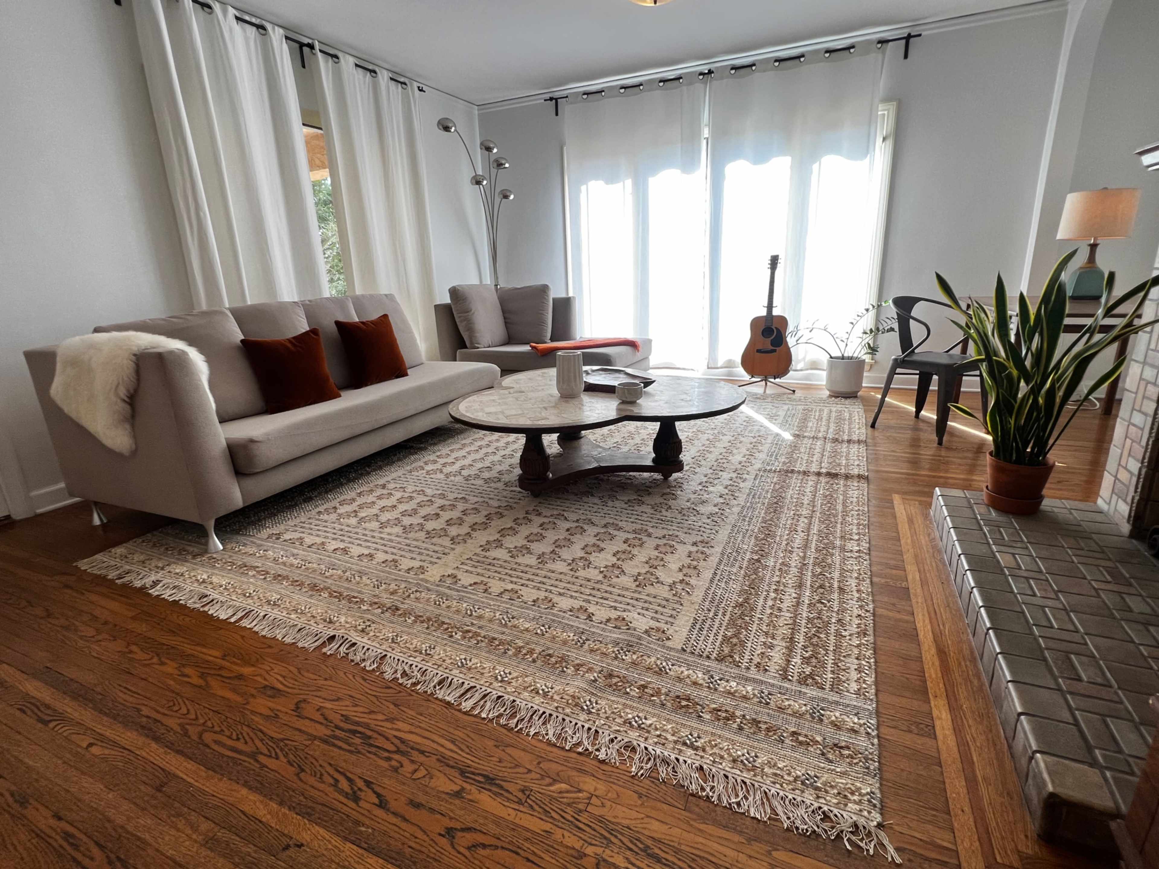 A light-filled living room with a sofa, a round coffee table, a guitar, and plants, all situated on a patterned area rug.
