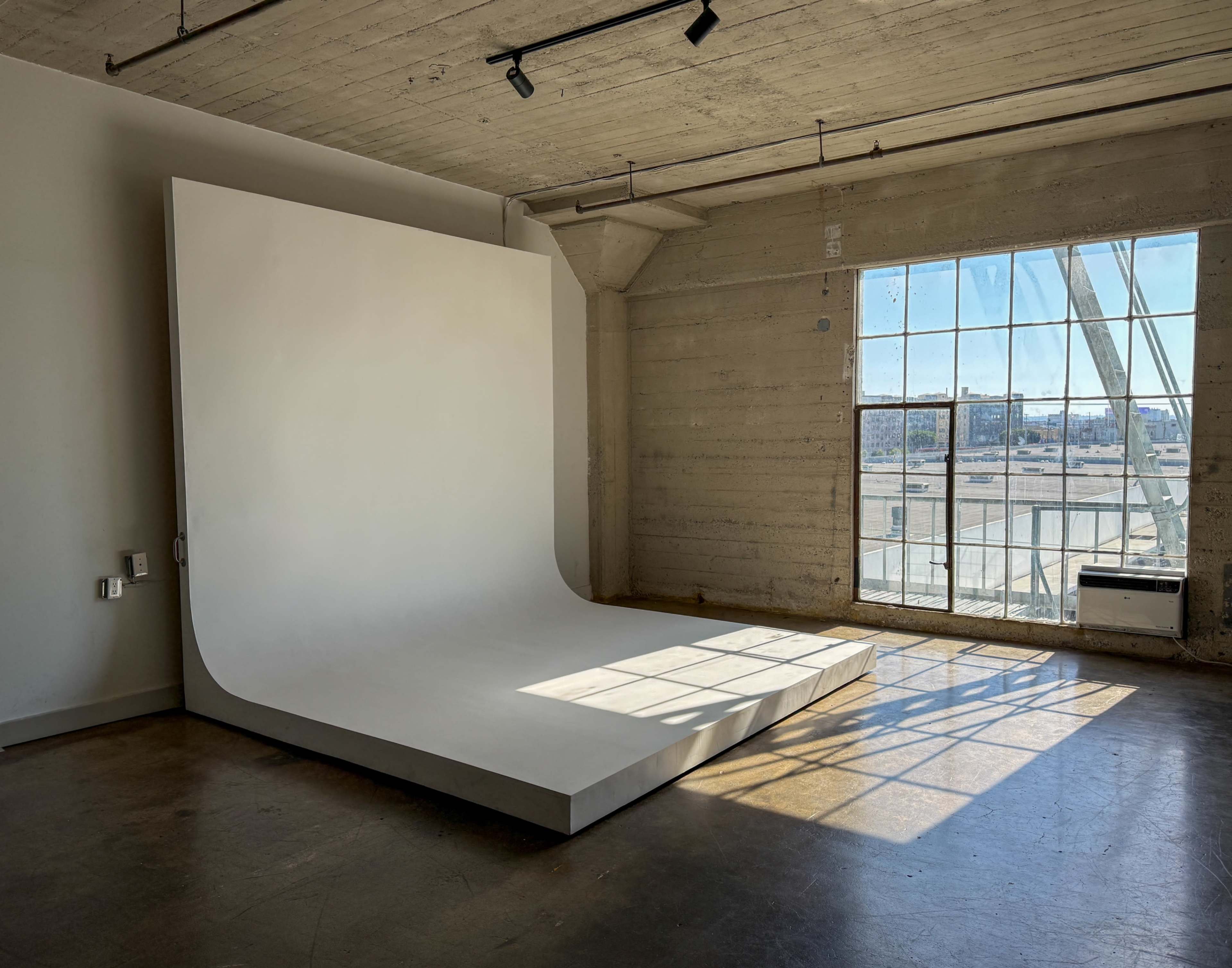 The image shows a minimalistic photo studio with a large white backdrop and a window allowing natural light to enter.