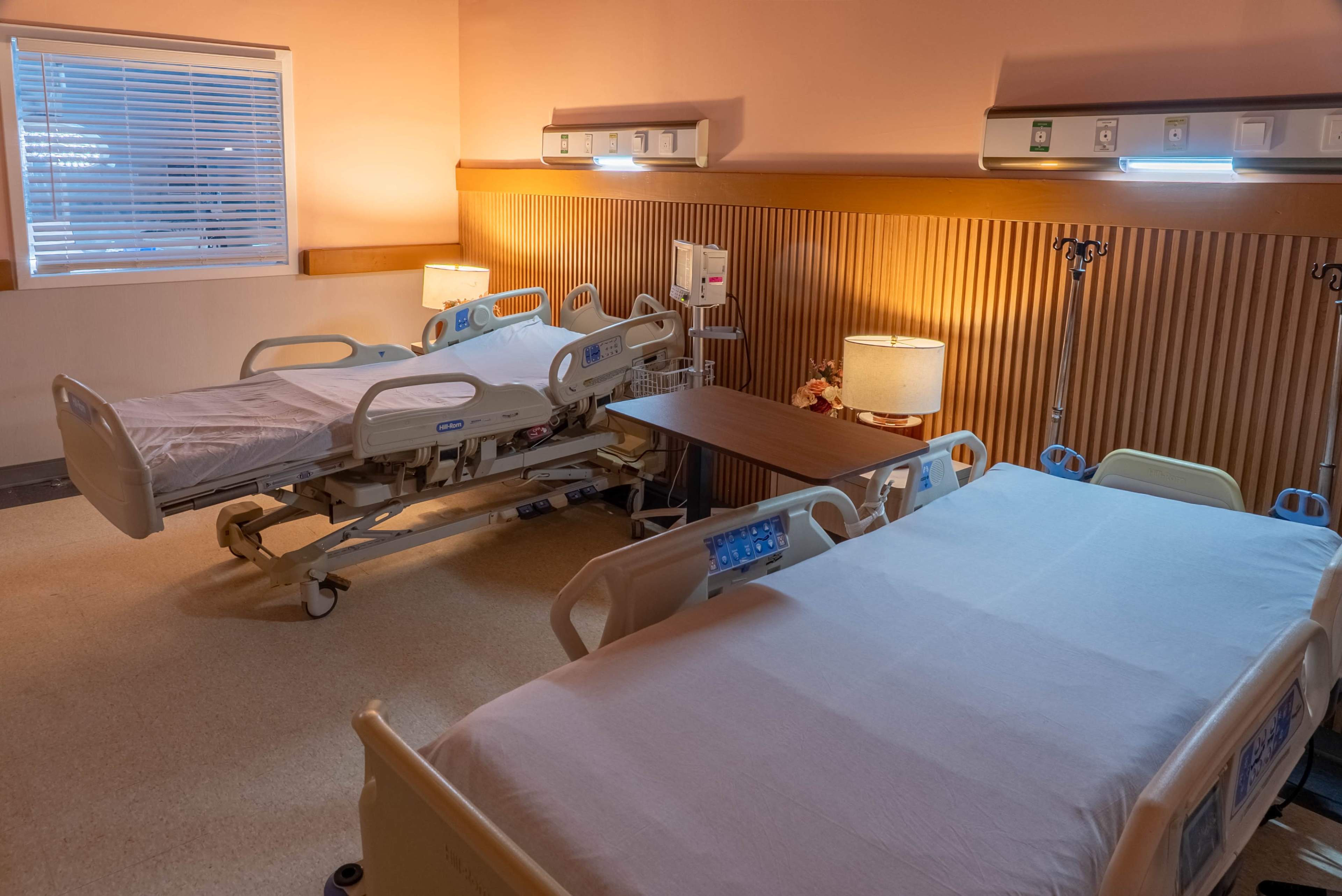 The image shows a hospital room with two beds, a bedside table, and a lamp, accompanied by a window letting in natural light.