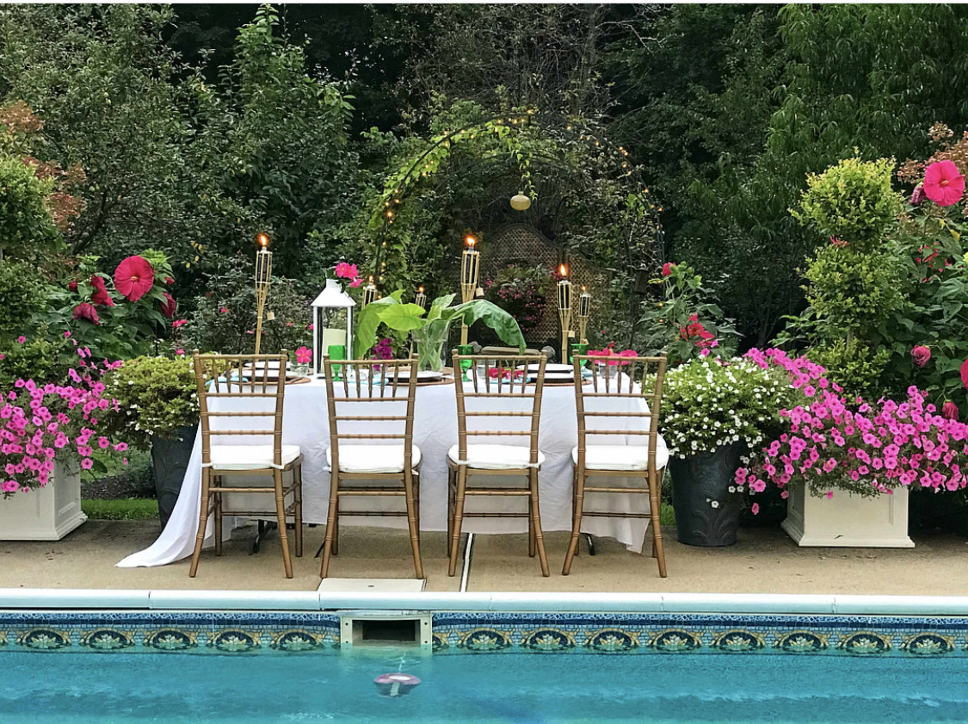 A beautifully set dining table with gold chairs is arranged by a swimming pool surrounded by vibrant flowers.