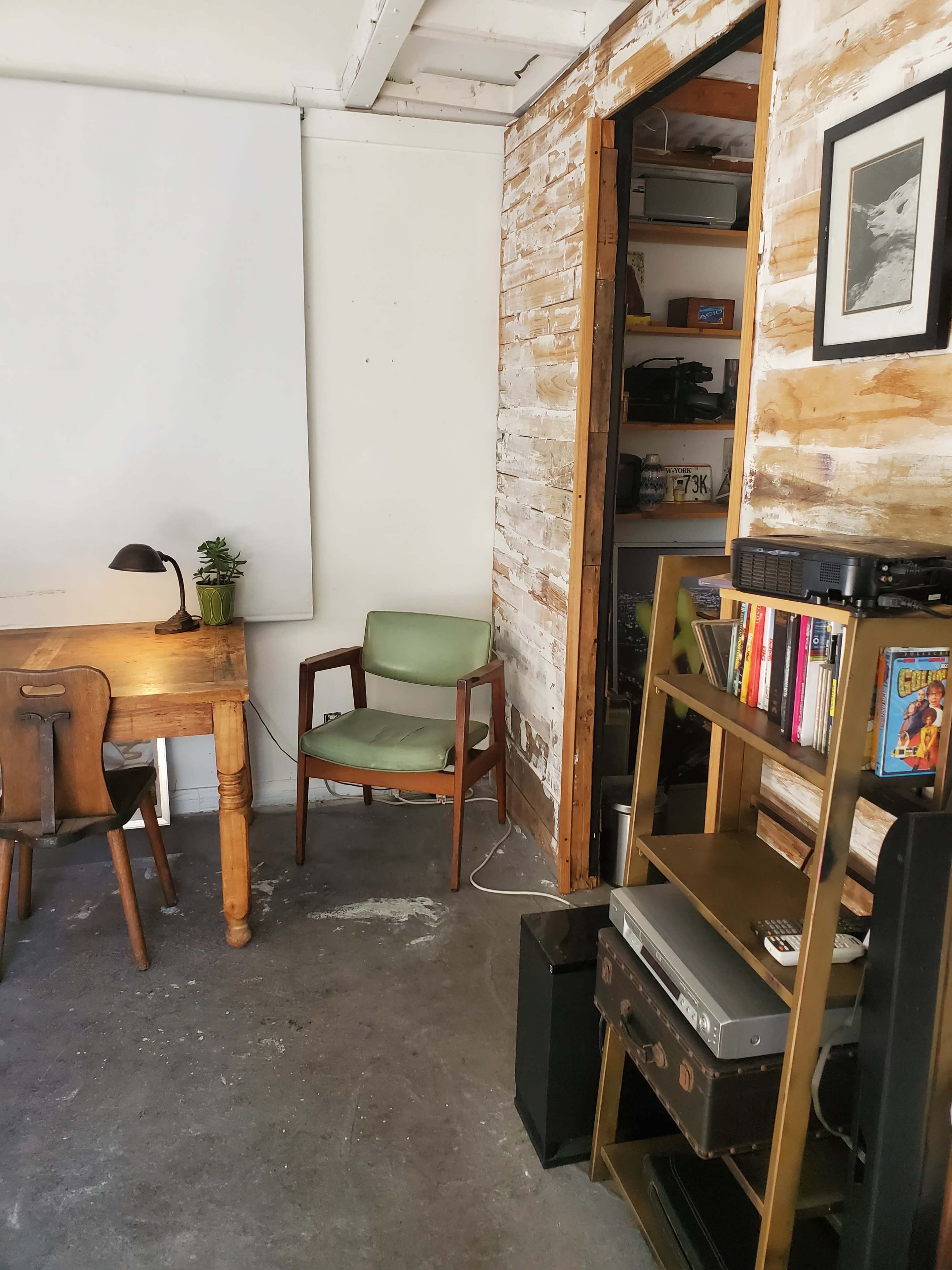 The image shows a small, minimalist workspace featuring a wooden desk with a lamp, a green chair, and a wall with exposed wooden panels and a bookshelf.