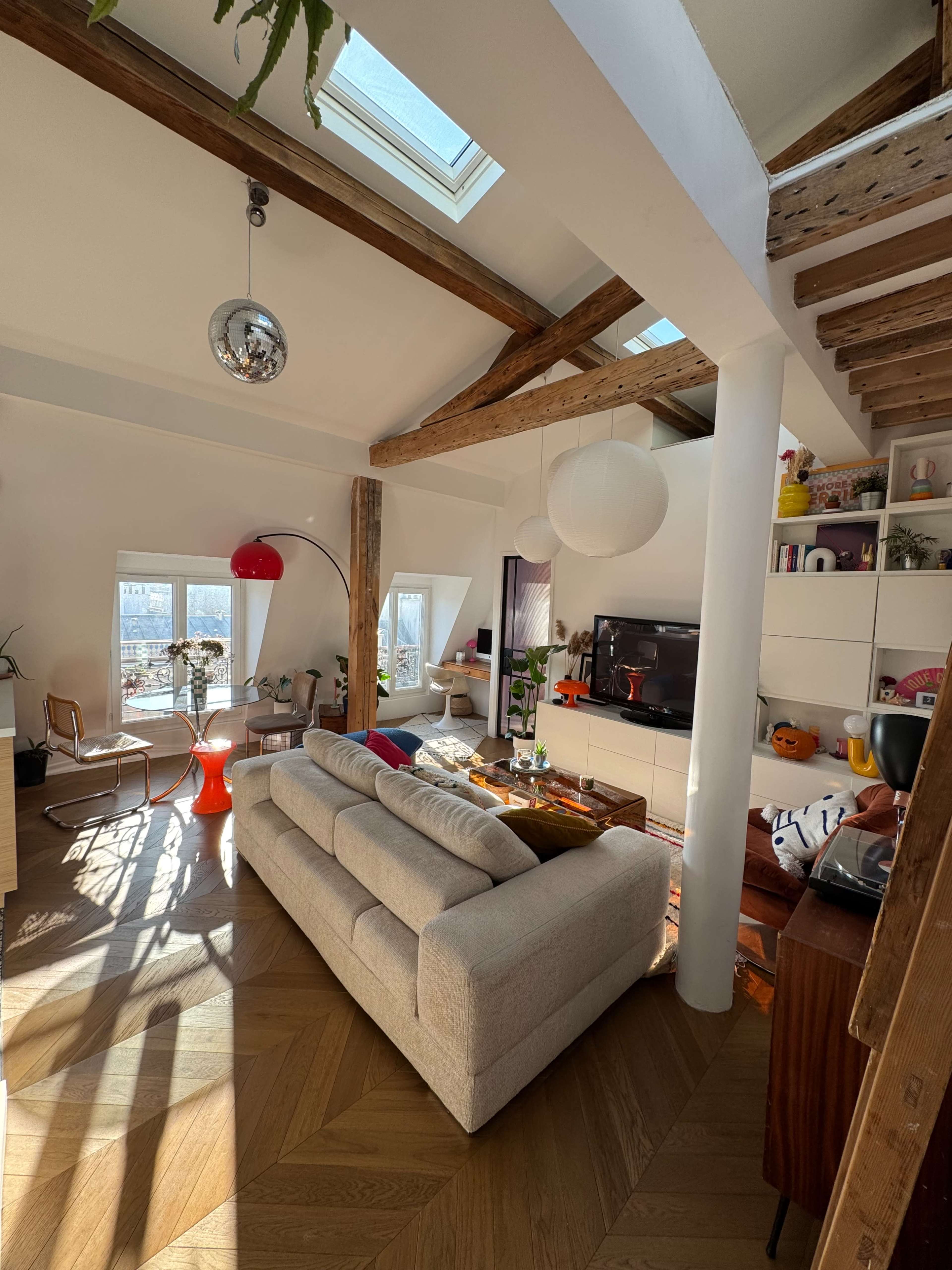 The image shows a modern living room with a light-colored sofa, wooden beams, and a skylight, featuring a coffee table and various decorative items.