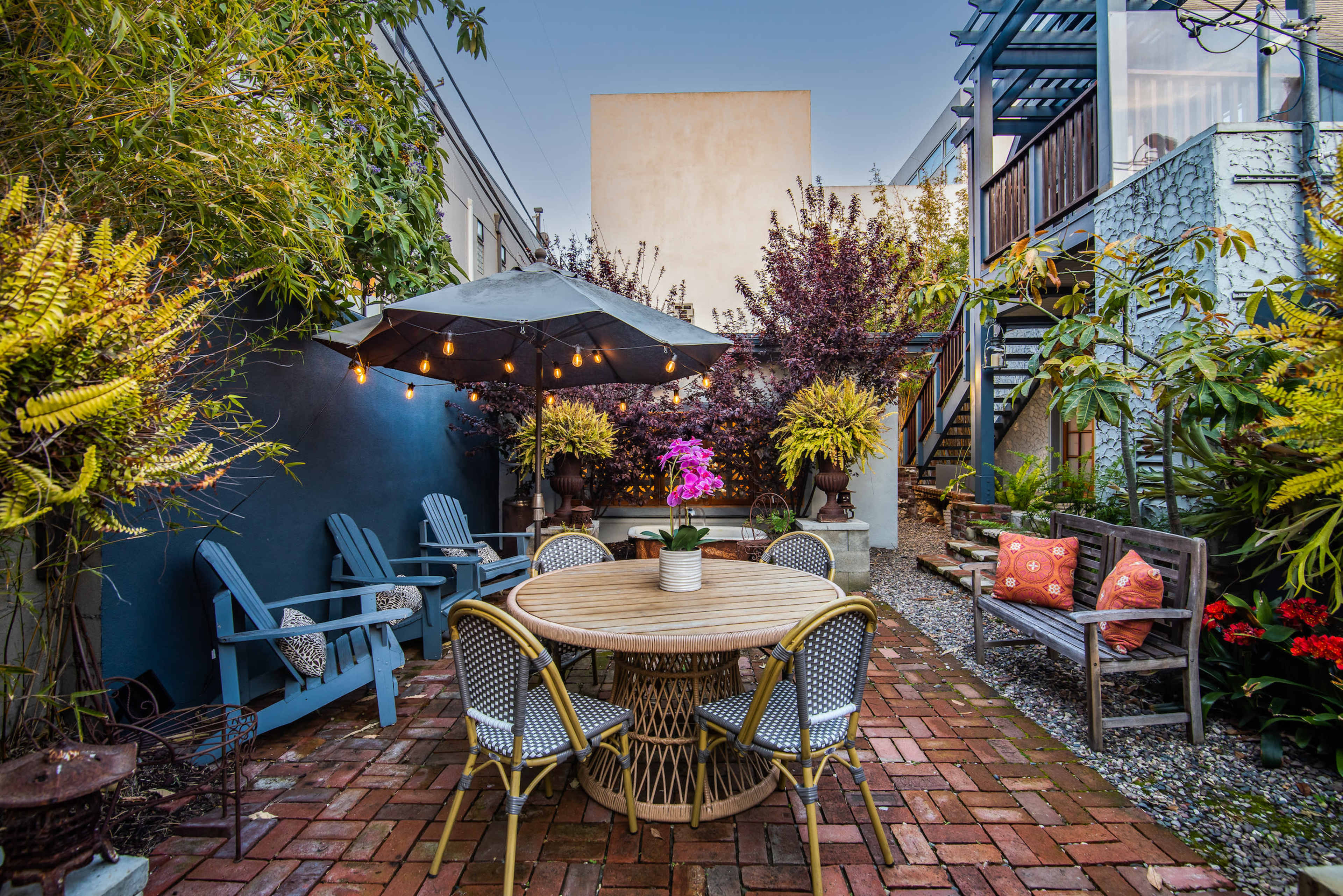 A cozy outdoor garden space featuring a round dining table surrounded by chairs, an umbrella for shade, and lush greenery with colorful plants and flowers.