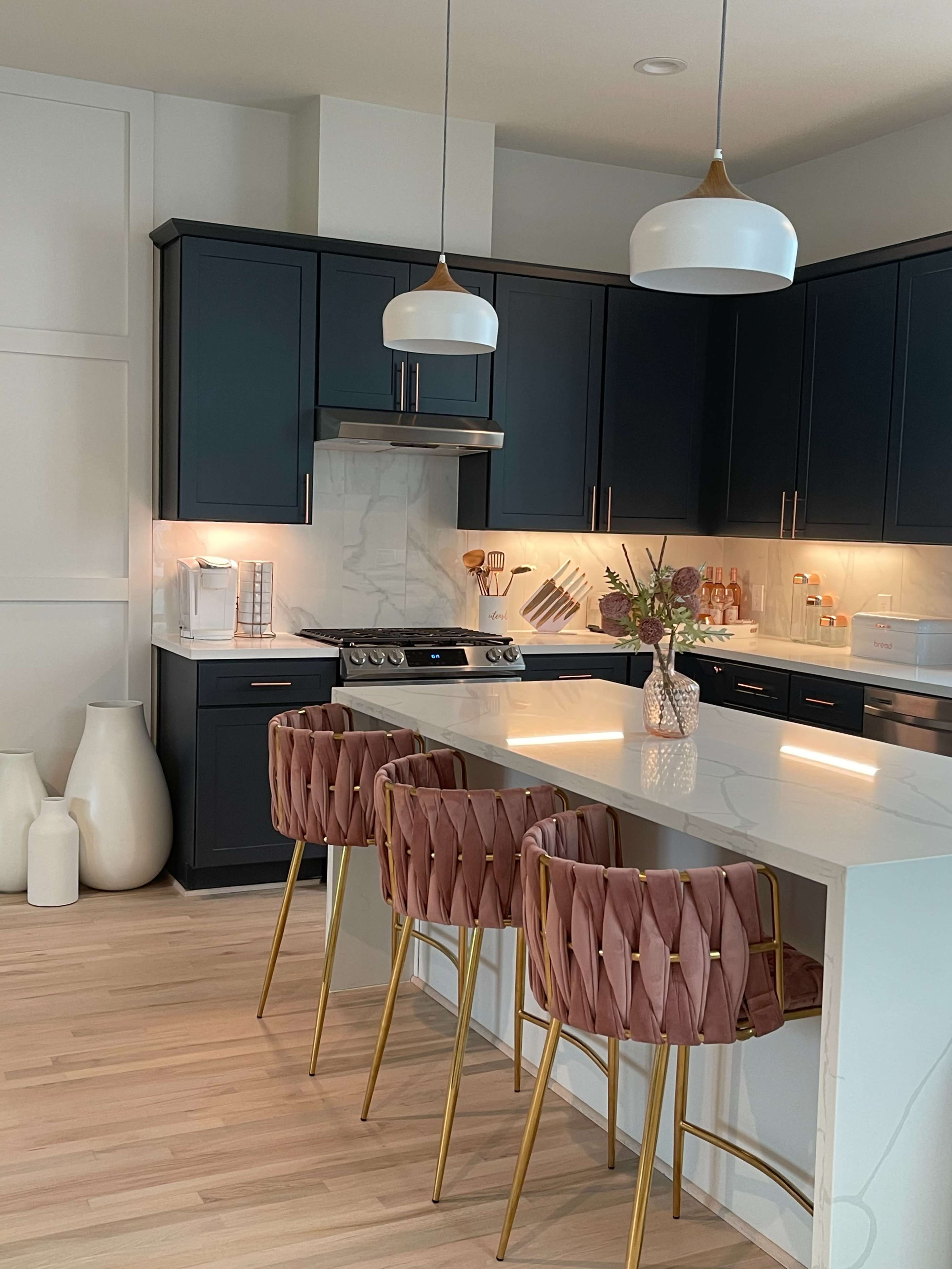 A modern kitchen with dark cabinetry, white countertops, and three pink and gold barstools at a central island.