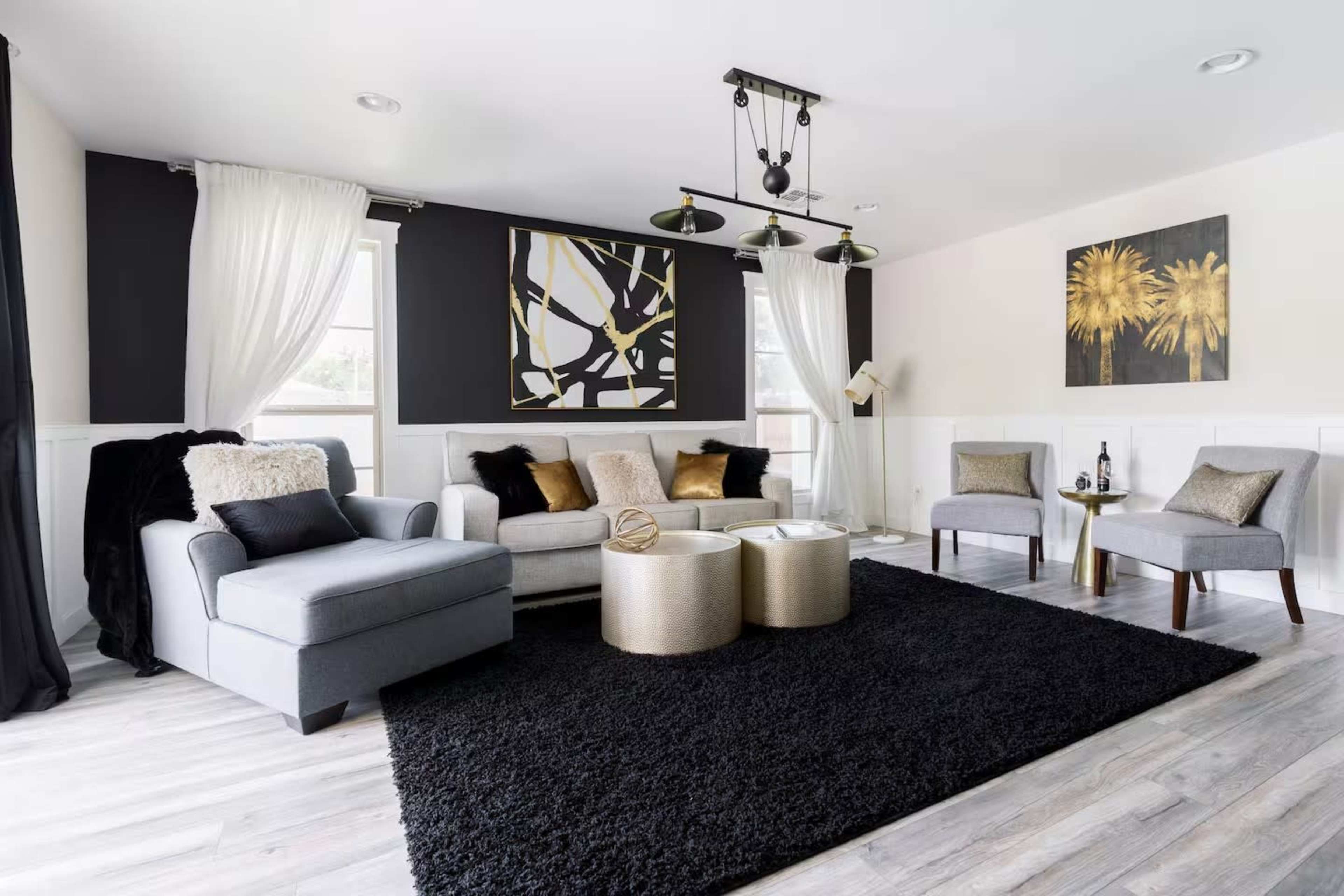 The living room features a gray sectional sofa, gold accent tables, and a black area rug, complemented by modern art and large windows with sheer curtains.
