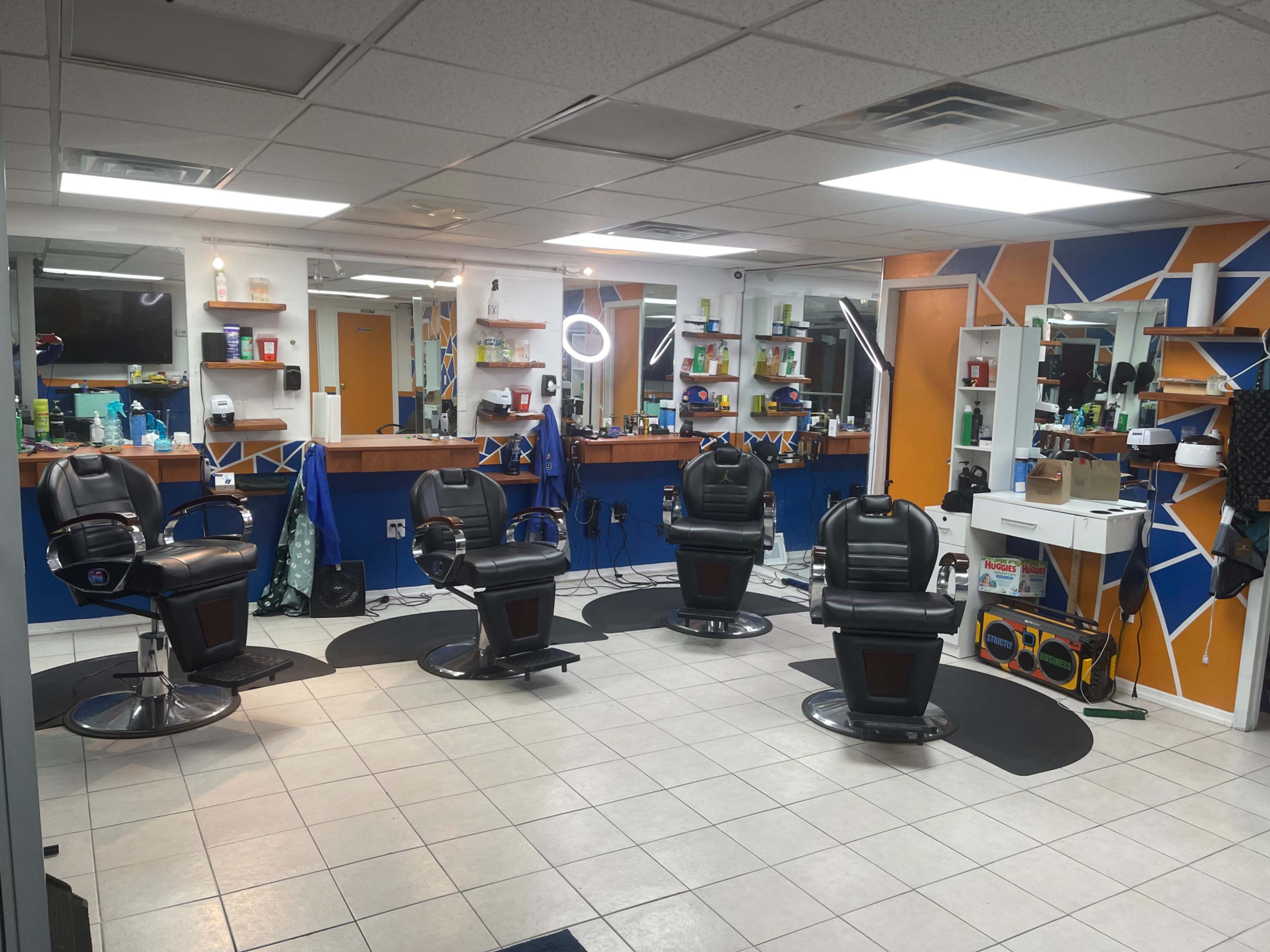 The image shows a modern barber shop with four black barber chairs arranged around a brightly colored wall, equipped with mirrors and various hair care products.