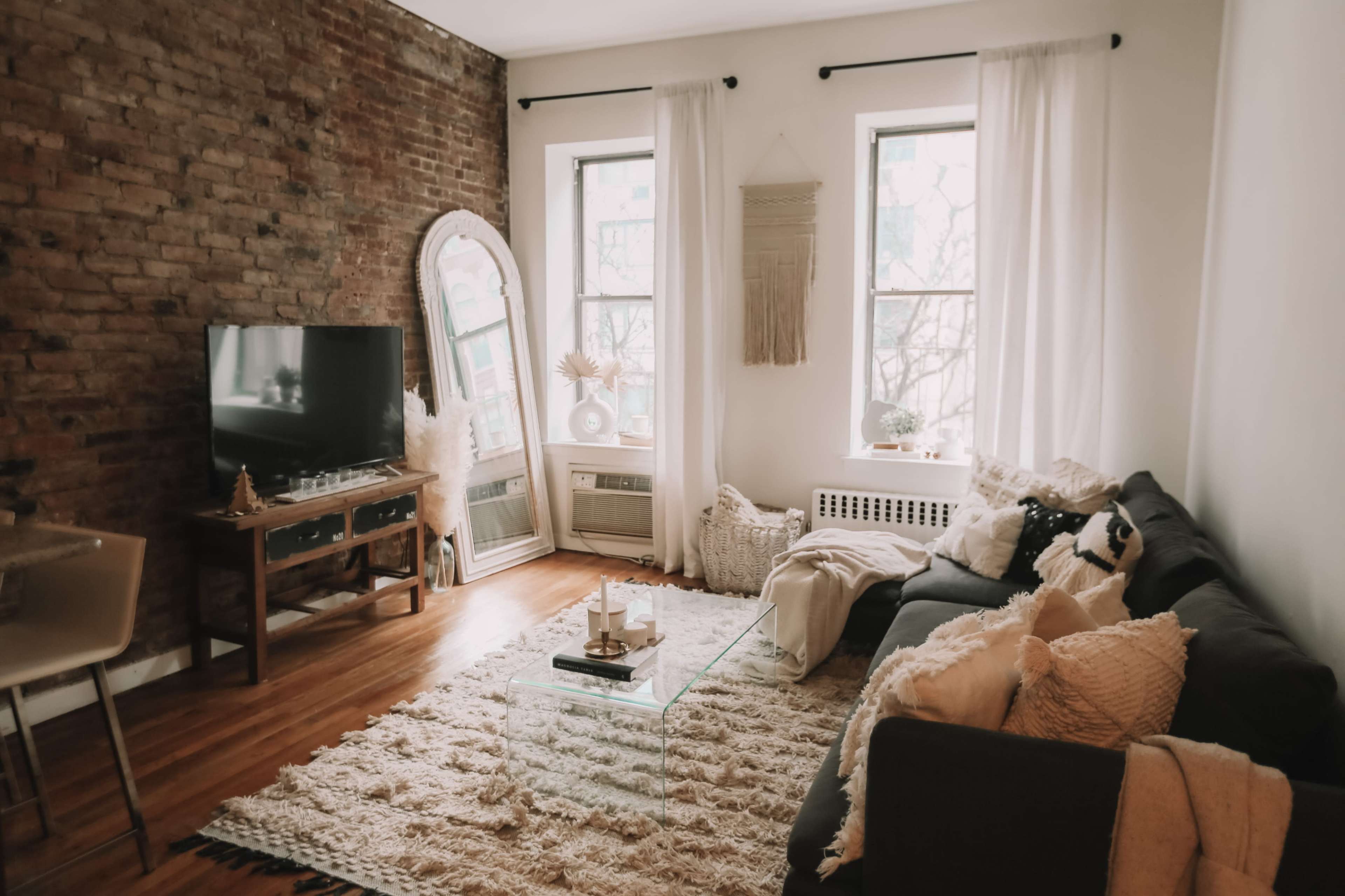 A cozy living room with exposed brick walls, large windows, a gray sofa, a glass coffee table, and a television stand.