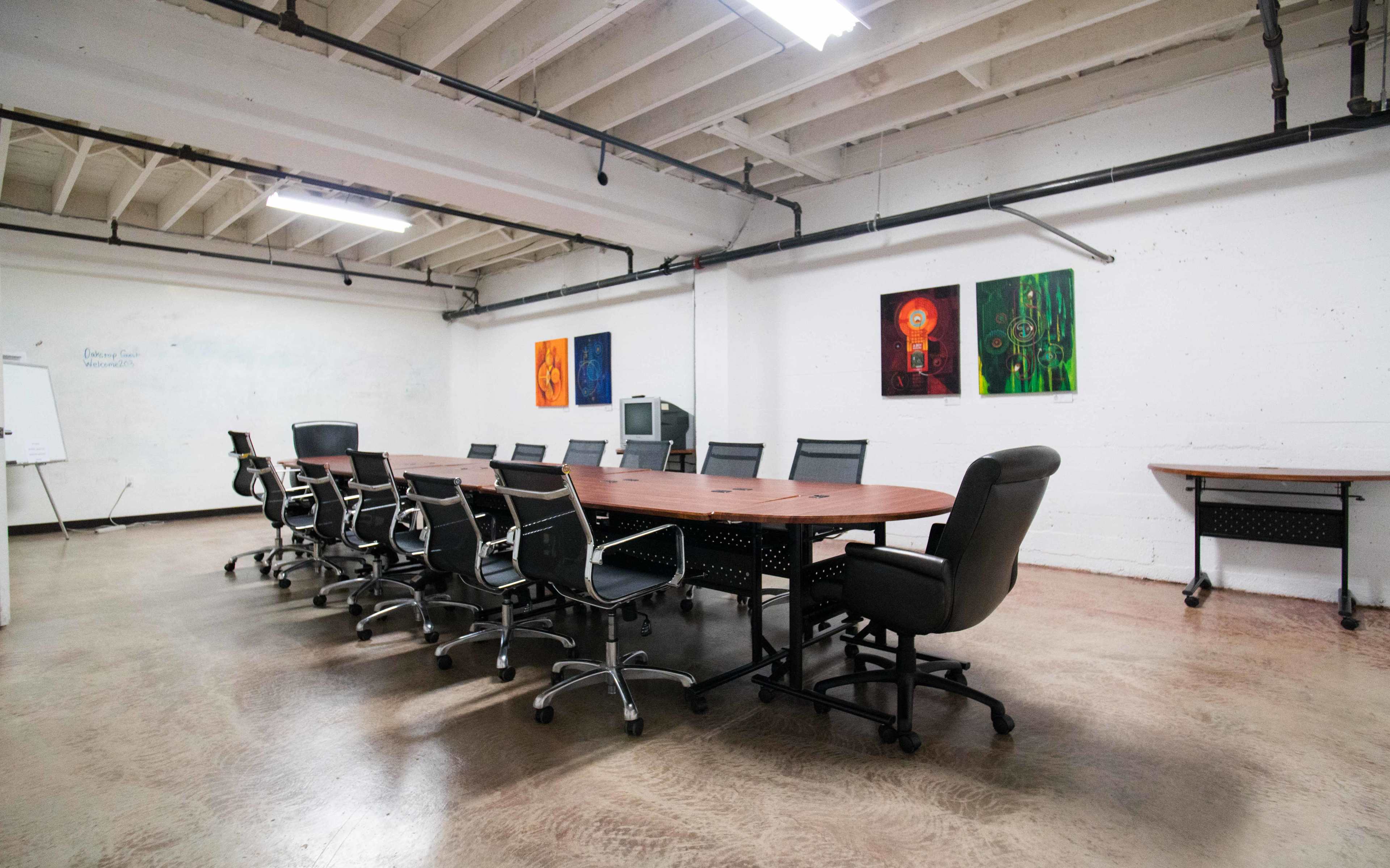 A long conference table surrounded by ergonomic chairs is positioned in a well-lit meeting room with colorful artwork on the walls.
