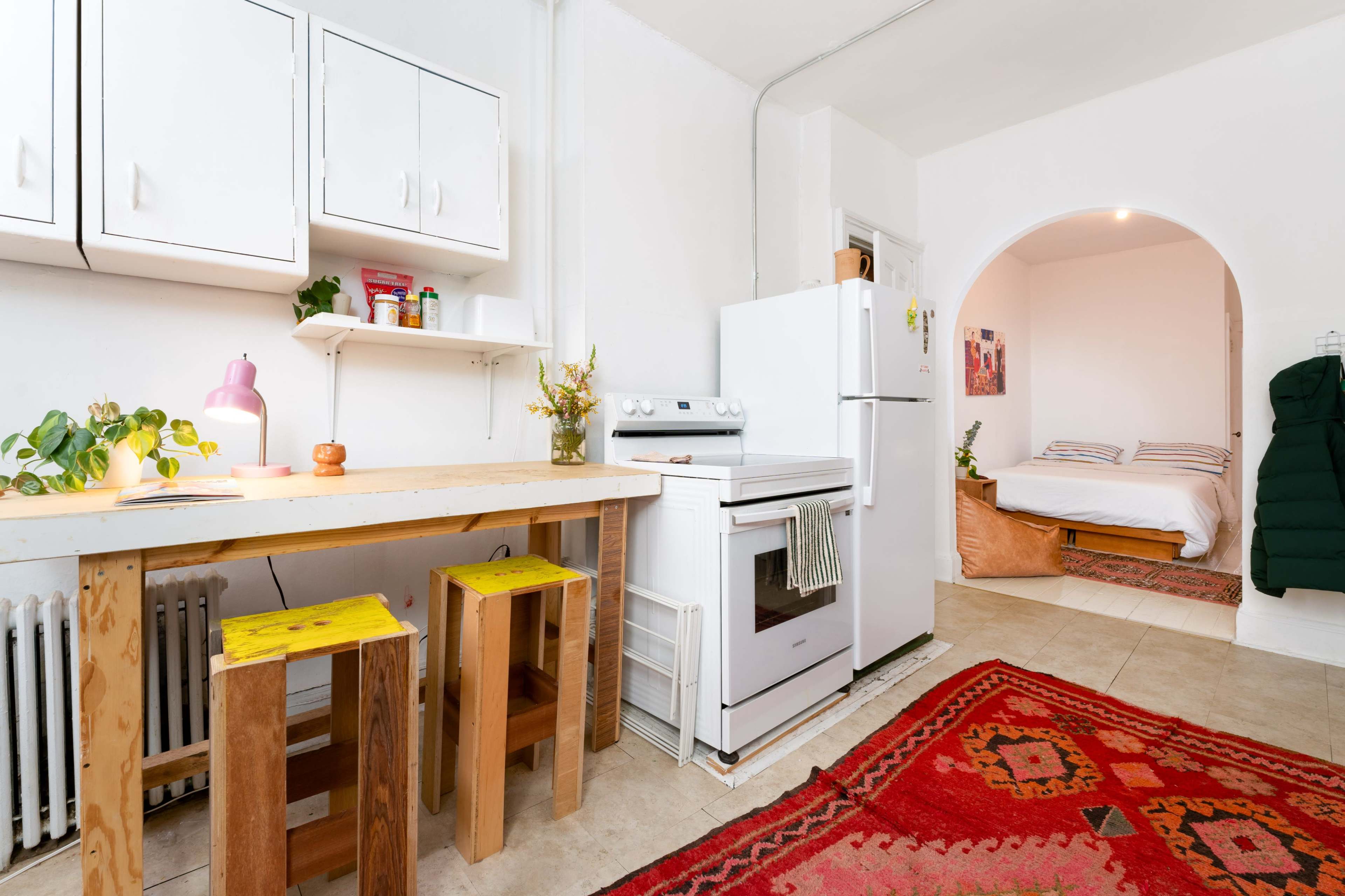 The image shows a small kitchen area with a wooden table and stools, a white refrigerator, an oven, and an archway leading to a bedroom.