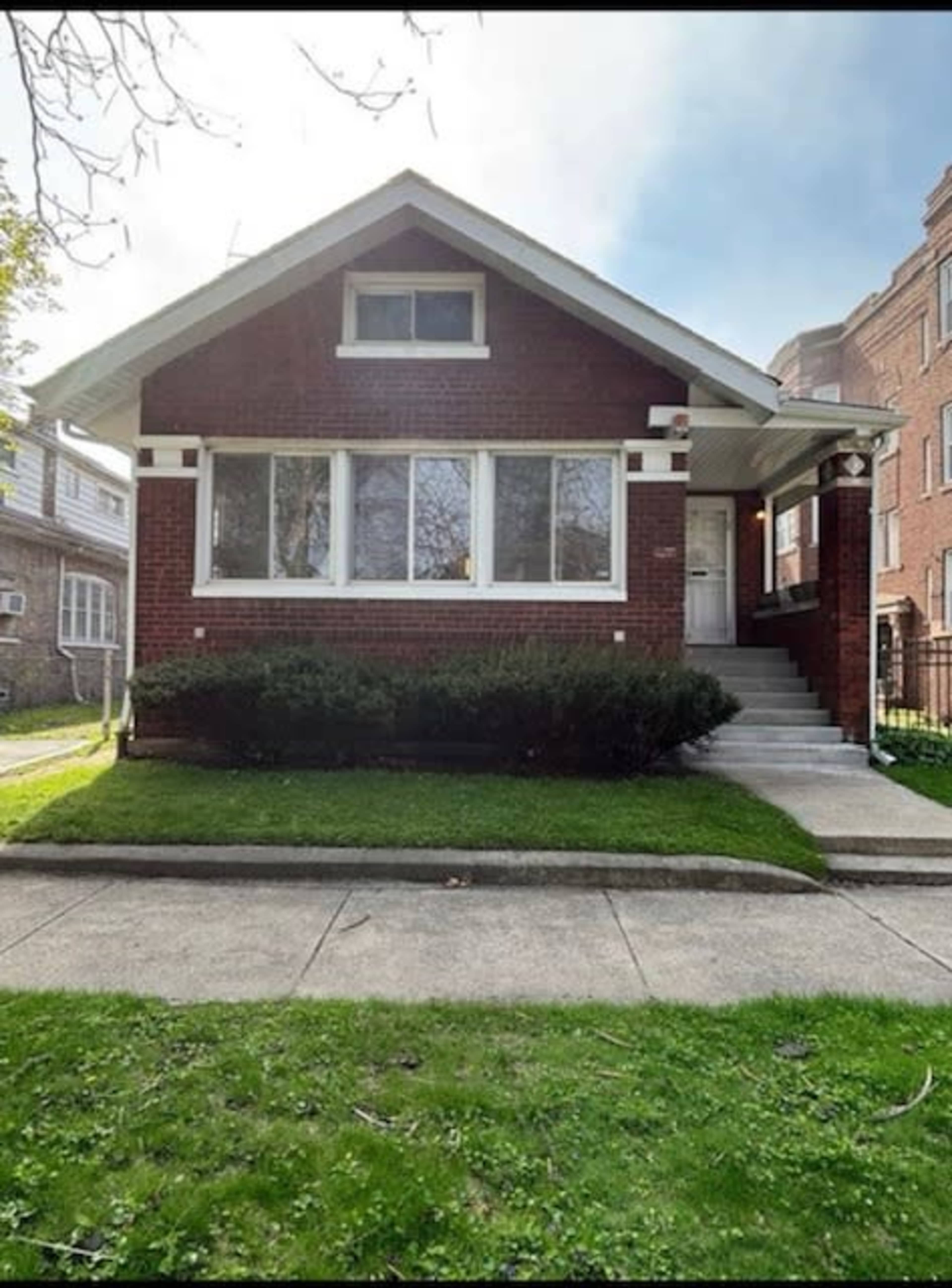 The image shows a red brick bungalow-style house with a front porch and large windows, situated on a grassy lawn.