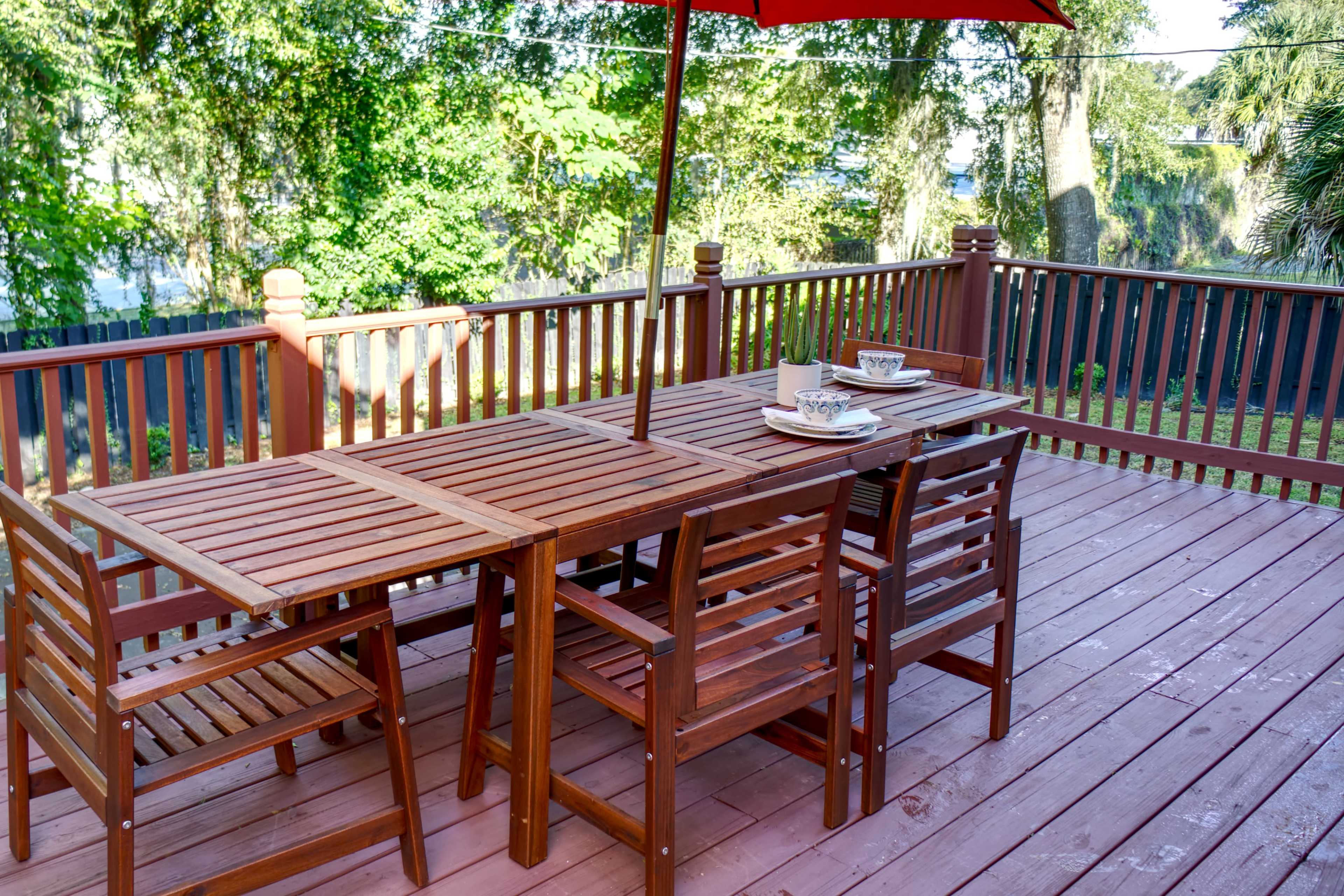 A wooden patio table and chairs set under a red umbrella is positioned on a deck surrounded by greenery.