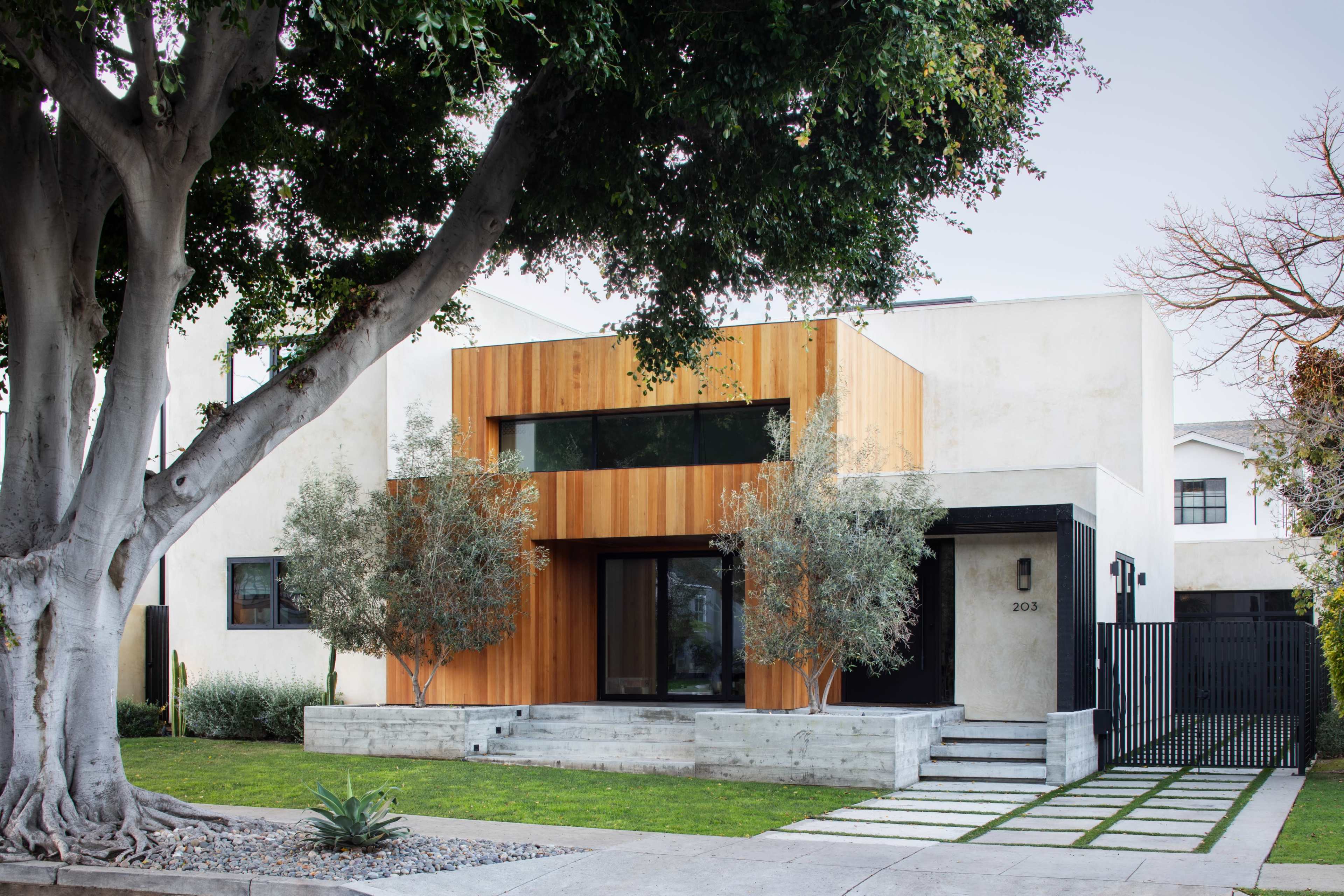 The image shows a modern two-story house featuring a wood-clad upper section, large windows, and a concrete lower level, surrounded by landscaped greenery and a mature tree.
