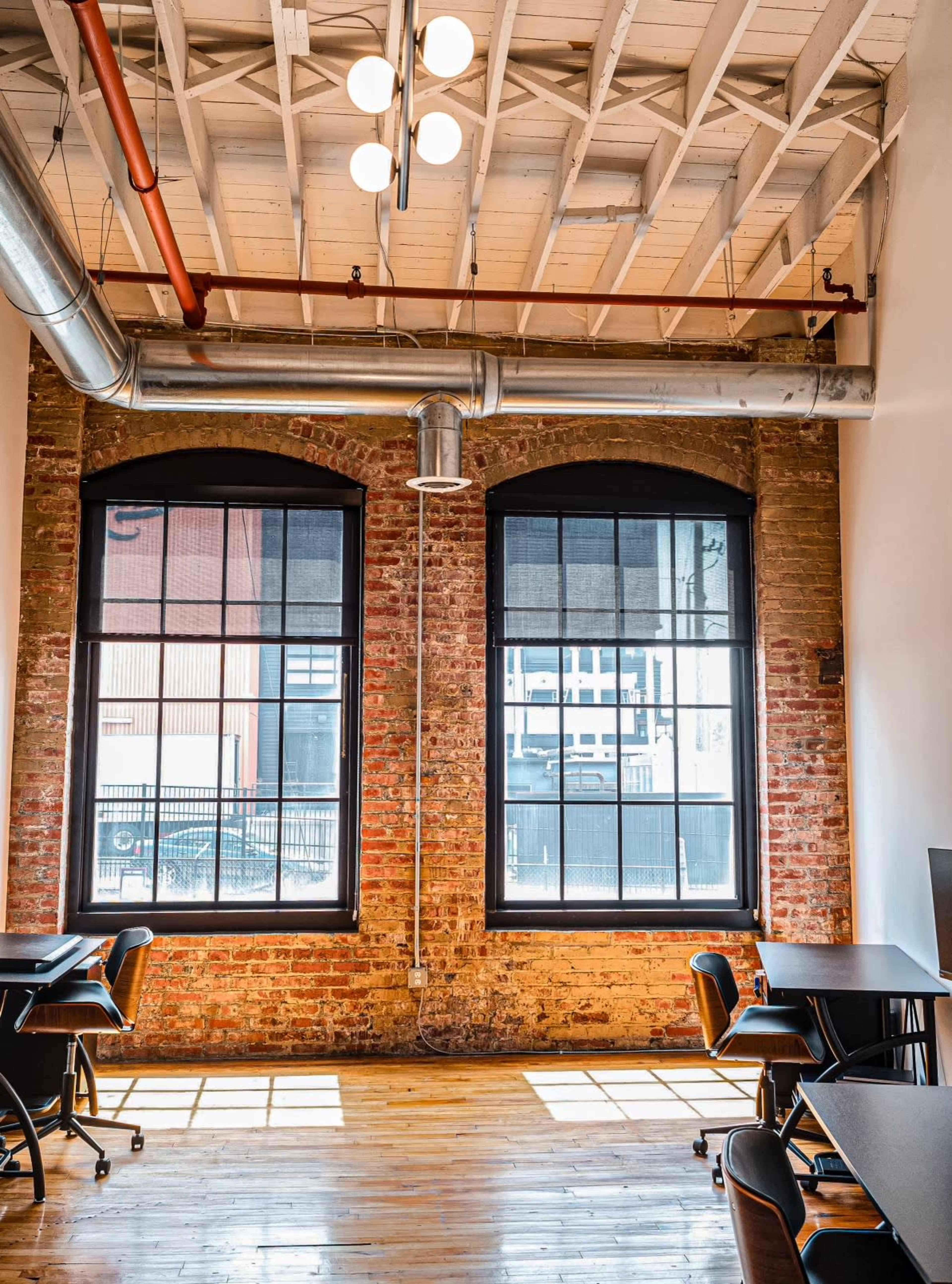 A modern office space with exposed brick walls, large windows, and wooden floors, featuring several desks arranged in the area.