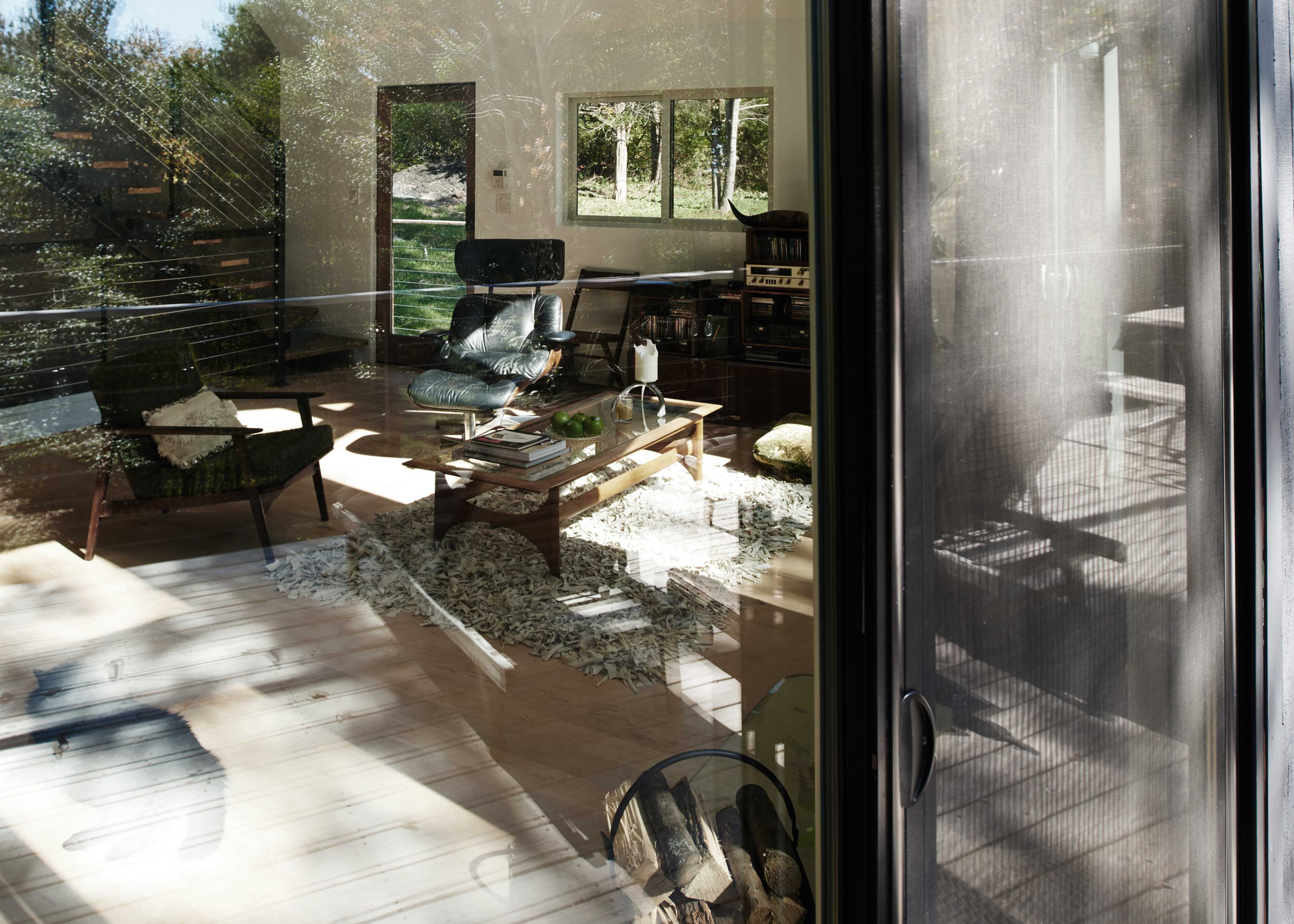 A modern living room features a coffee table surrounded by chairs, visible through large glass doors reflecting the outdoor greenery.
