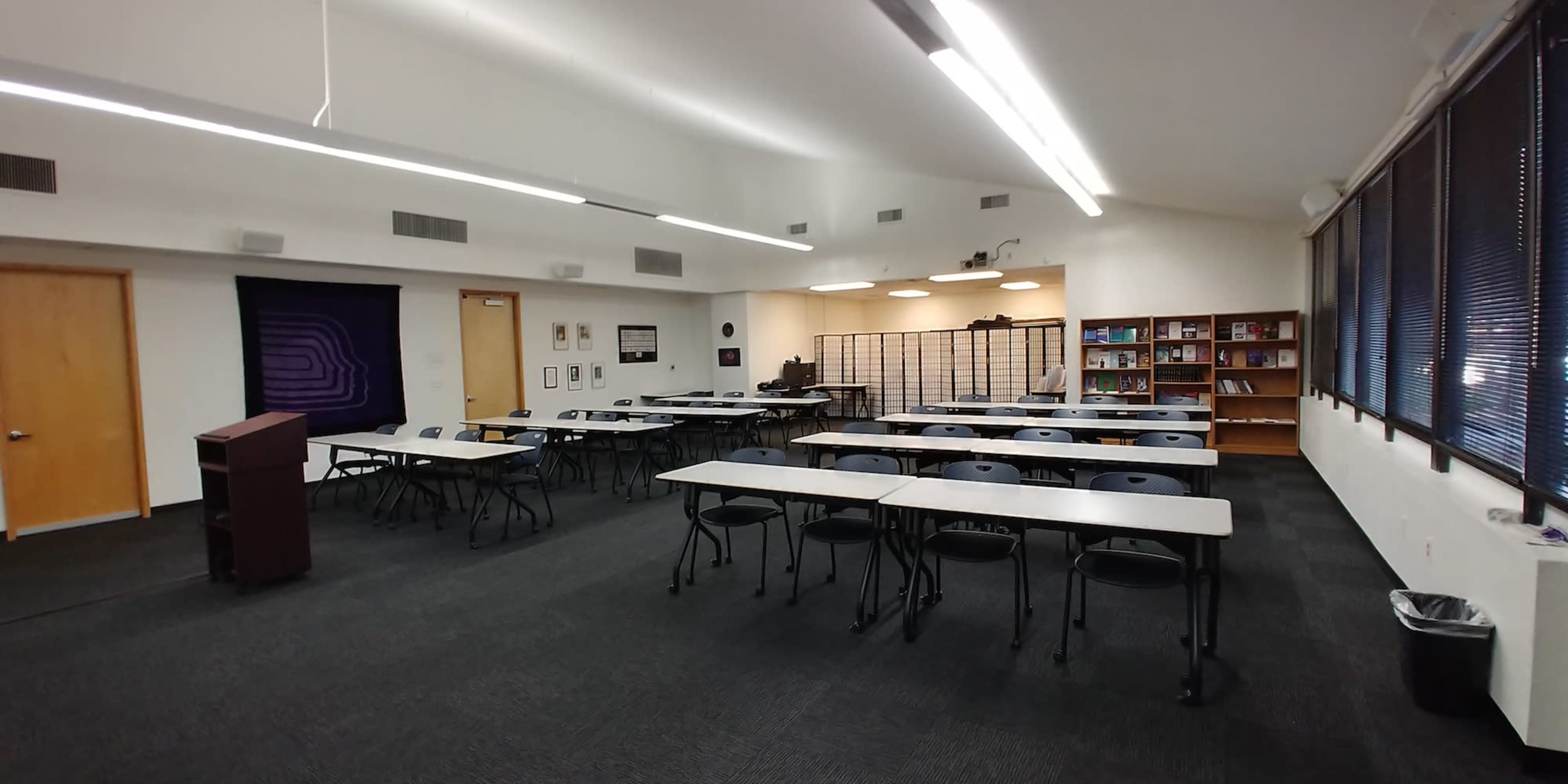A classroom setting with rows of tables and chairs, a podium at the front, and bookshelves along one wall.