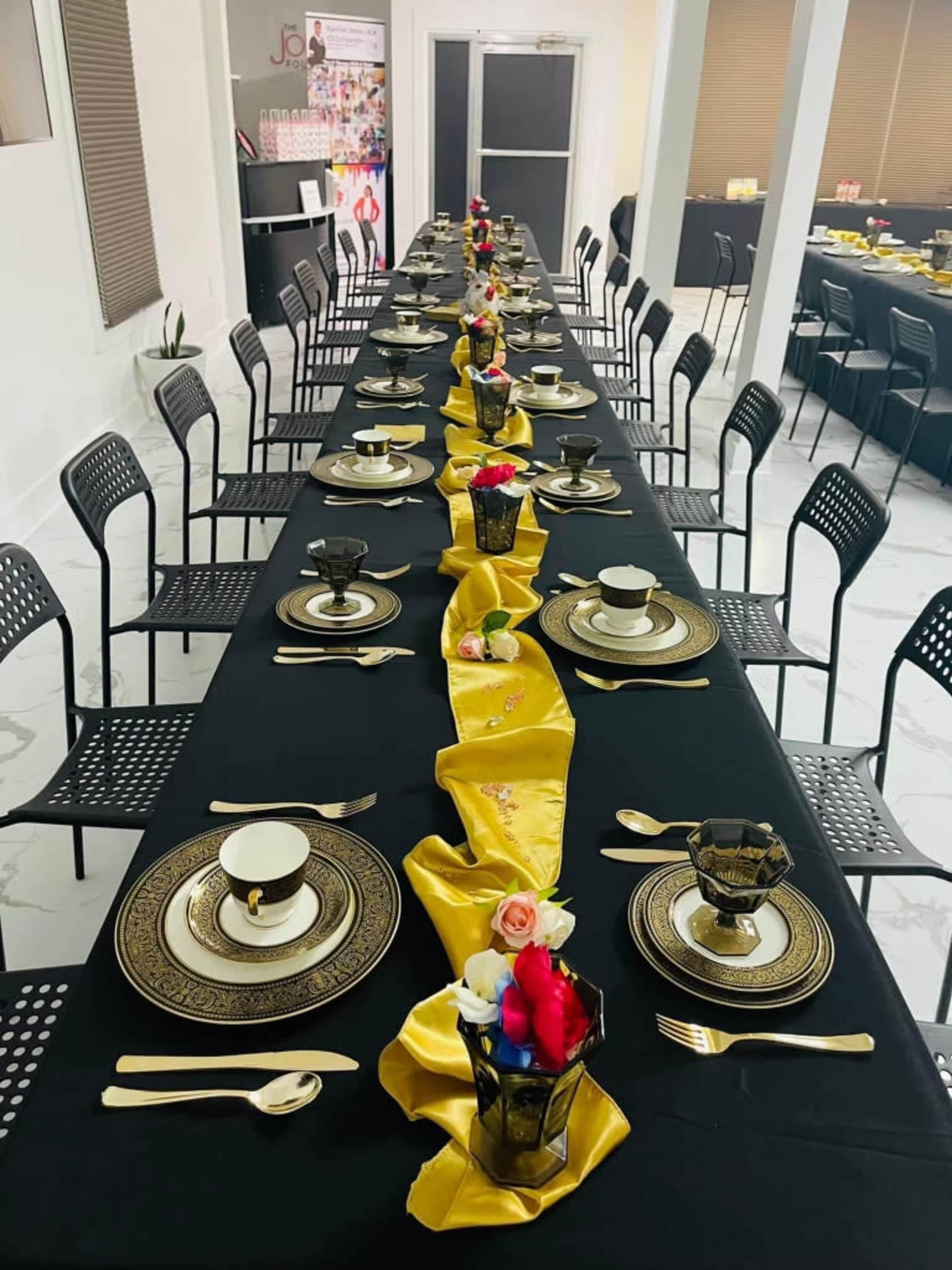 A long, elegantly set dining table is displayed with black tablecloths, gold accents, decorative tableware, and a center arrangement of flowers.