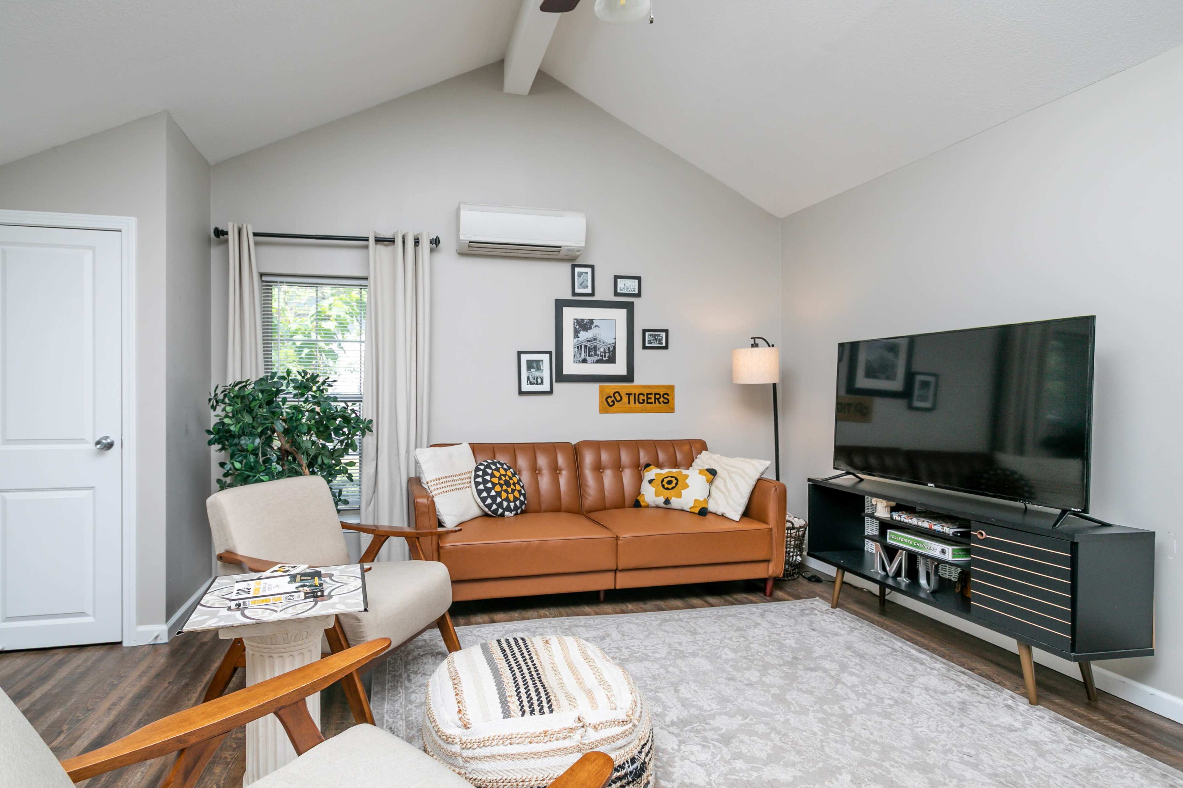 The living room features a tan leather sofa, a television on a stand, and a decorative rug, complemented by plants and wall art.