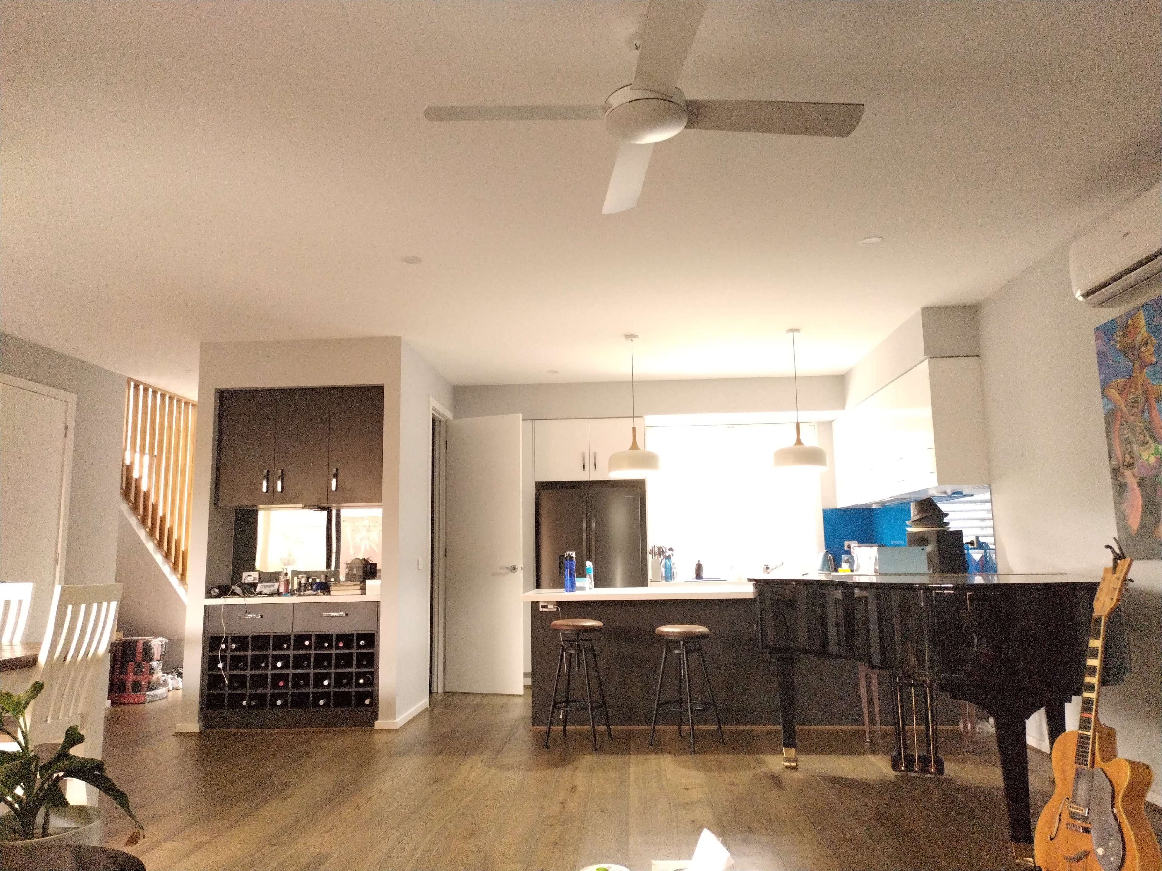The image shows a modern kitchen and living area featuring a piano, bar stools, and a wine rack, with a ceiling fan above.