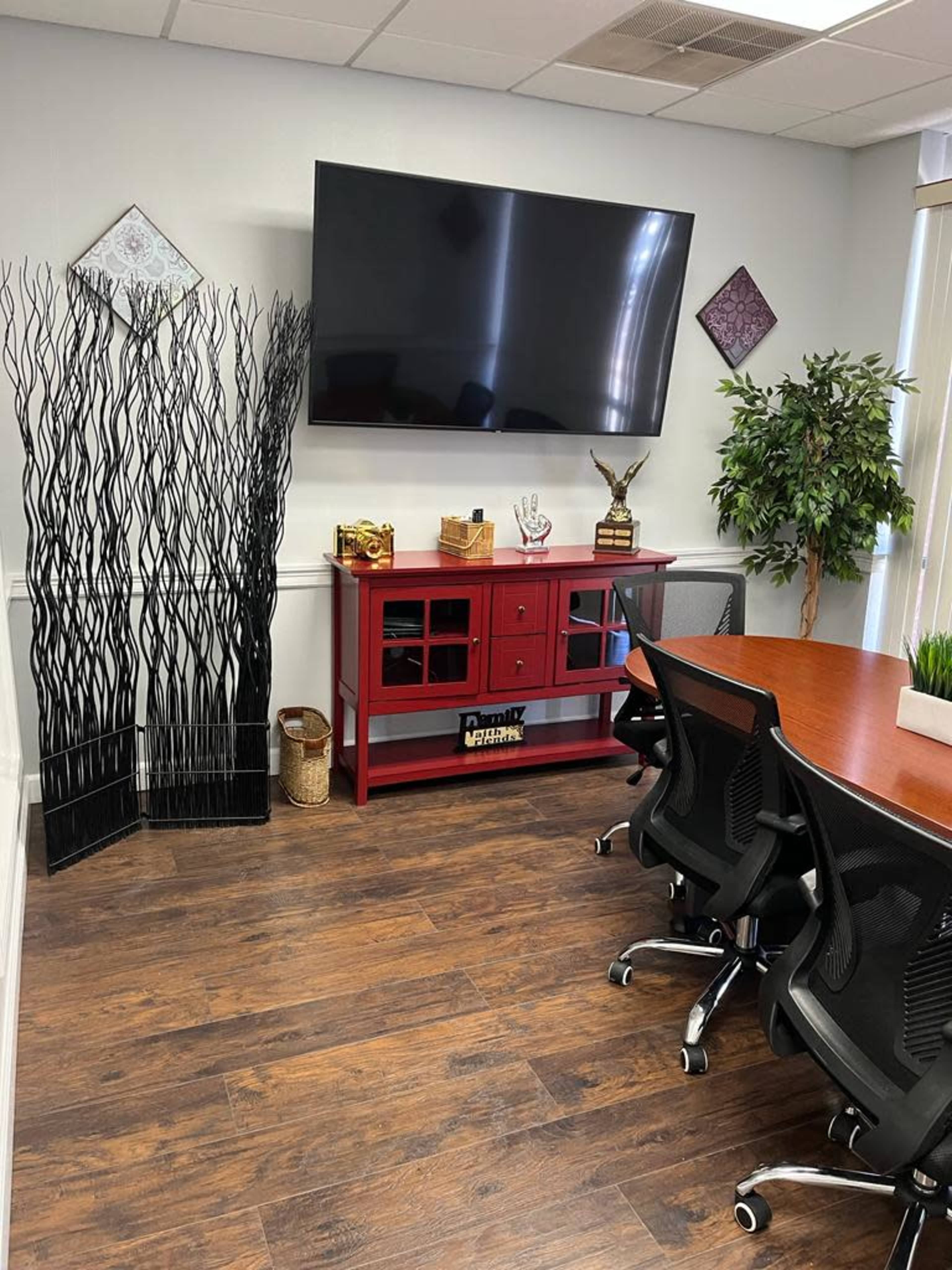 A modern office meeting room with a large flat-screen TV mounted on the wall, a red storage cabinet, a wooden conference table, and several black ergonomic chairs.