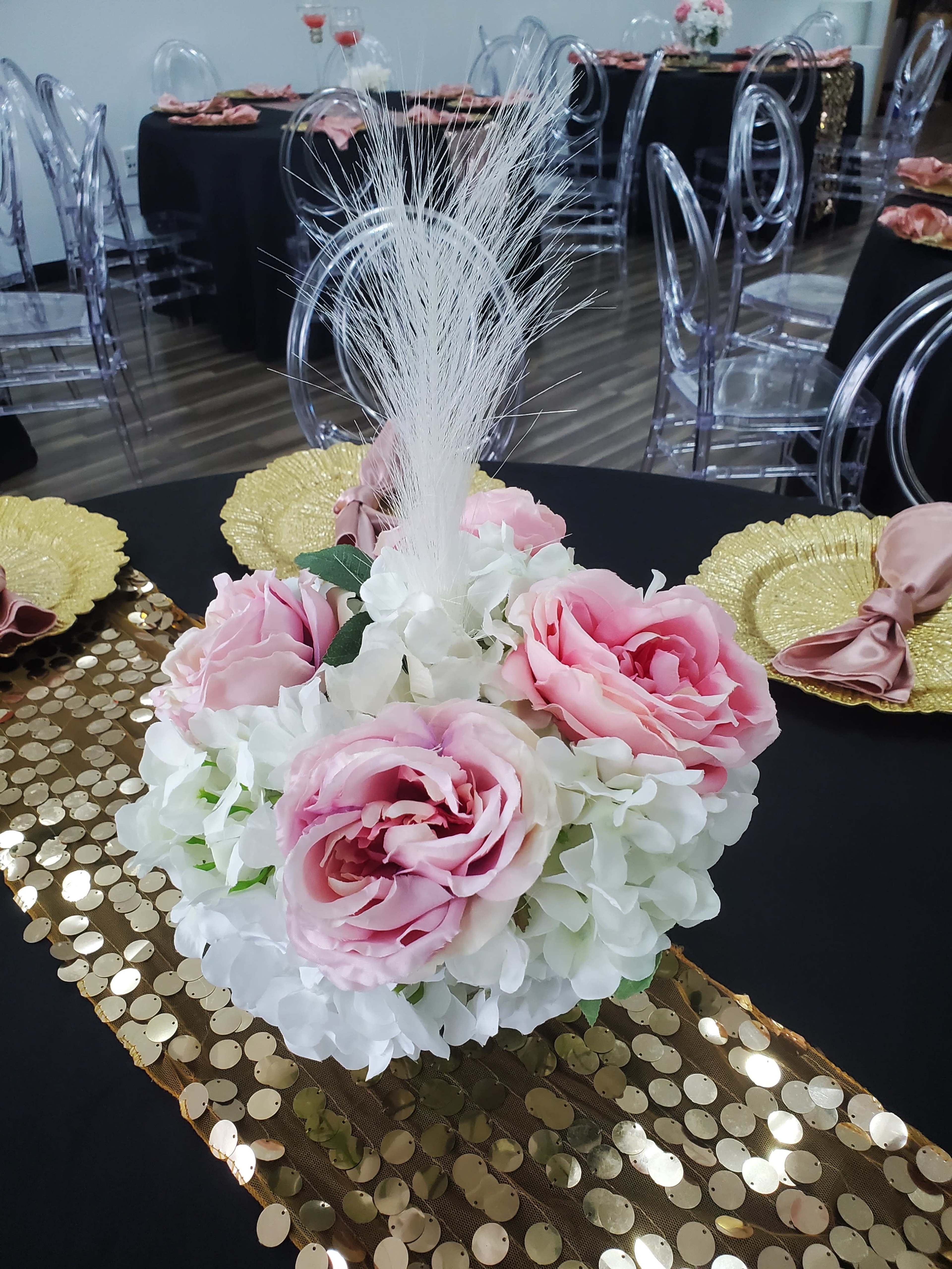 A floral centerpiece featuring pink roses and white hydrangeas is displayed on a table with a gold sequin runner and clear chairs in the background.