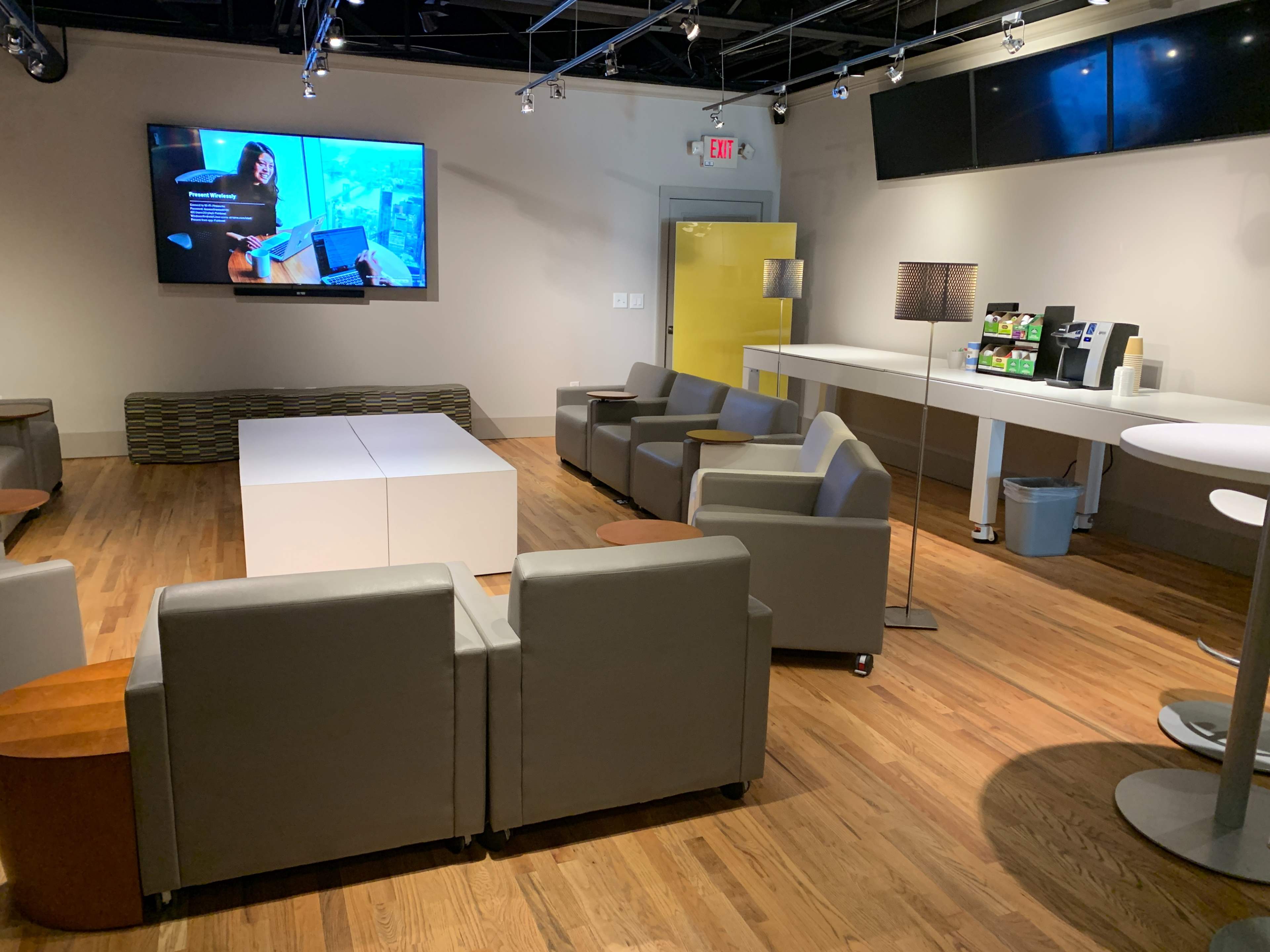 A modern lounge area with gray seating, a coffee station, and a large screen displaying content on a wall.
