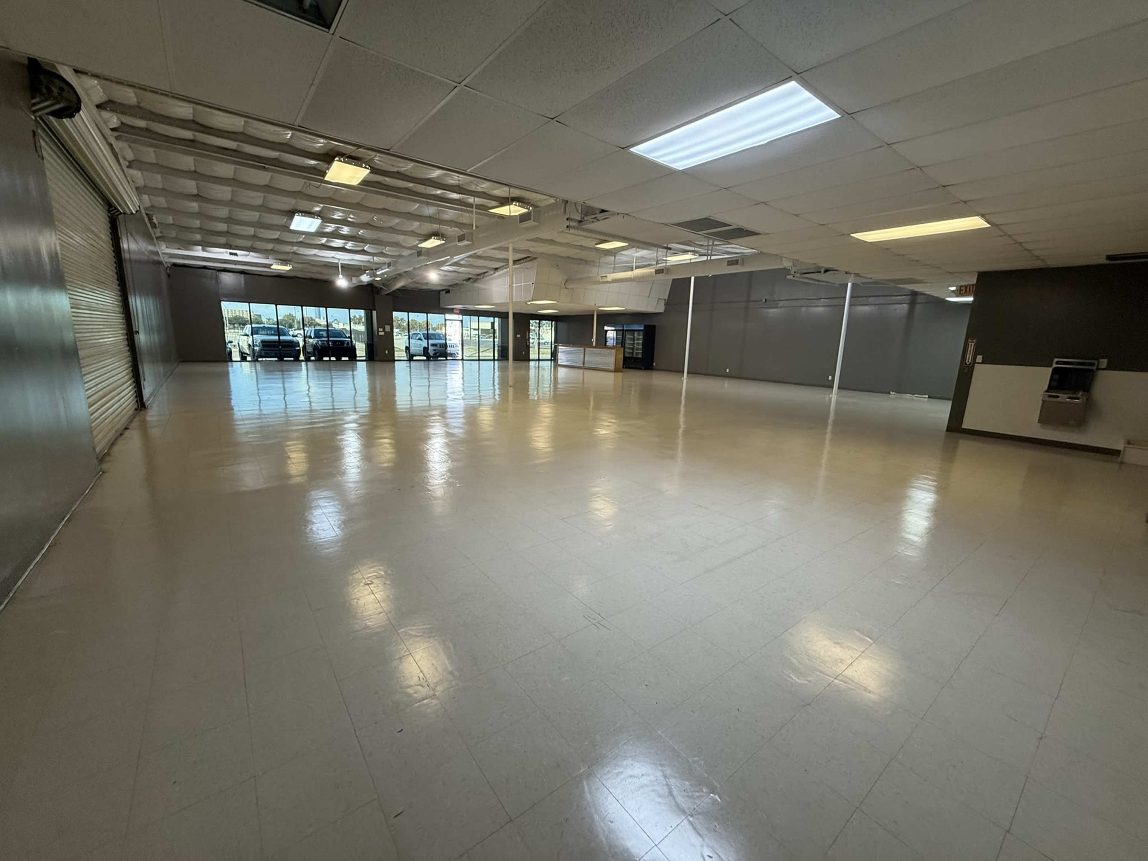 The image shows a large, empty commercial space with white tiled flooring and large windows along one wall.
