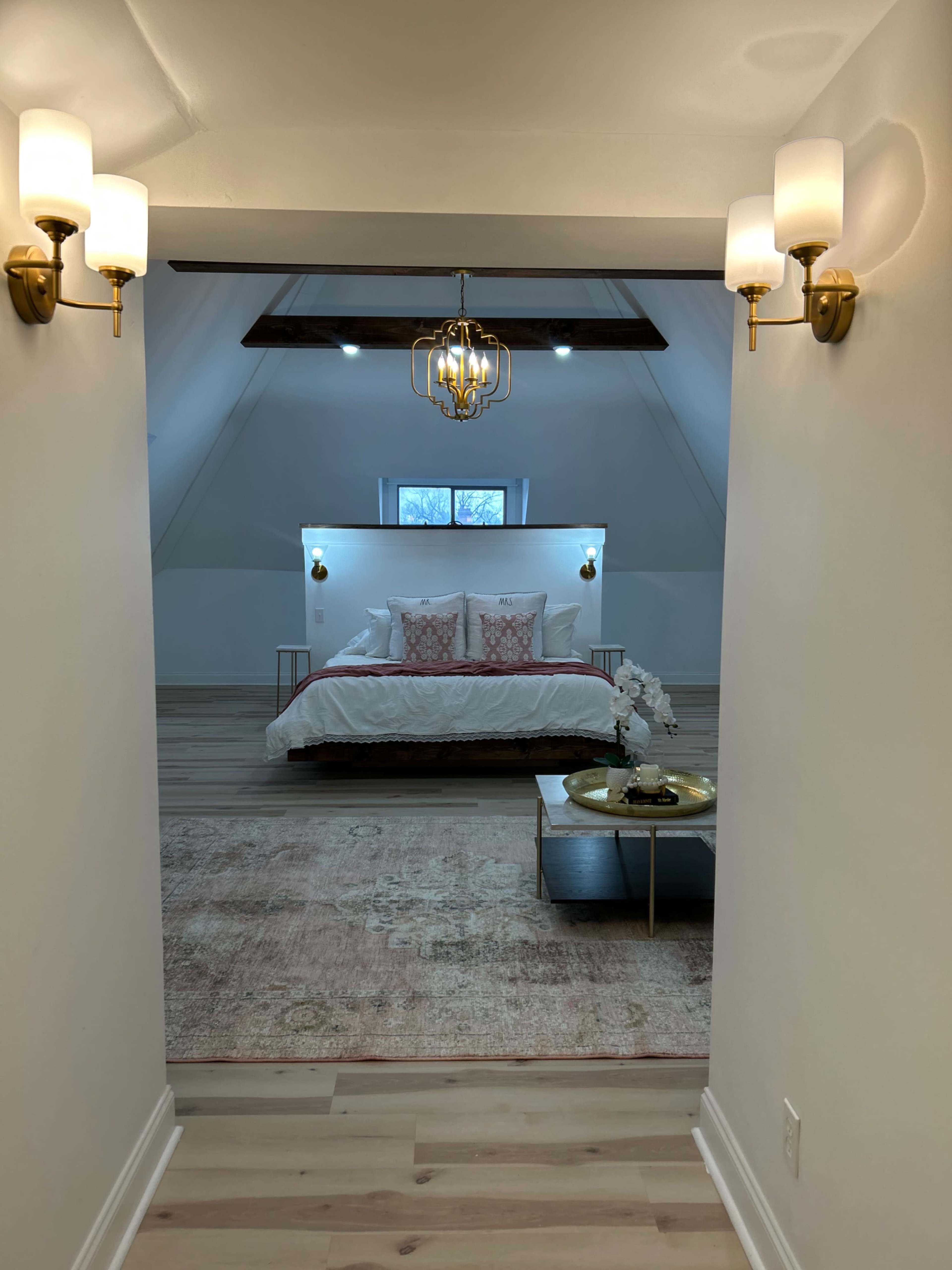 A well-lit attic bedroom features a bed with white bedding and decorative pillows, framed by sleek lighting fixtures and a wooden beam ceiling.