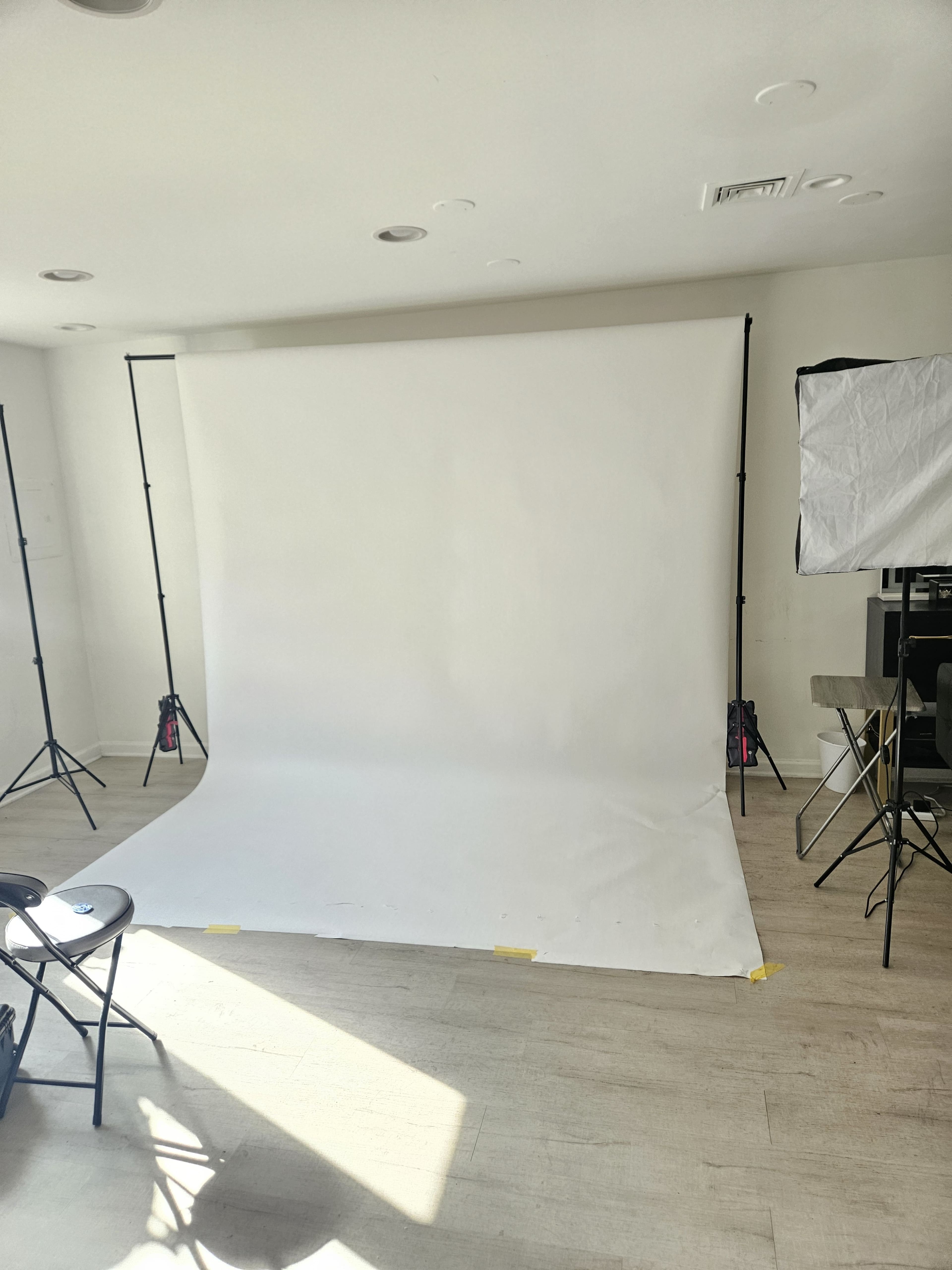 The image shows a photography studio setup with a white backdrop, lighting equipment, and black chairs arranged in the room.