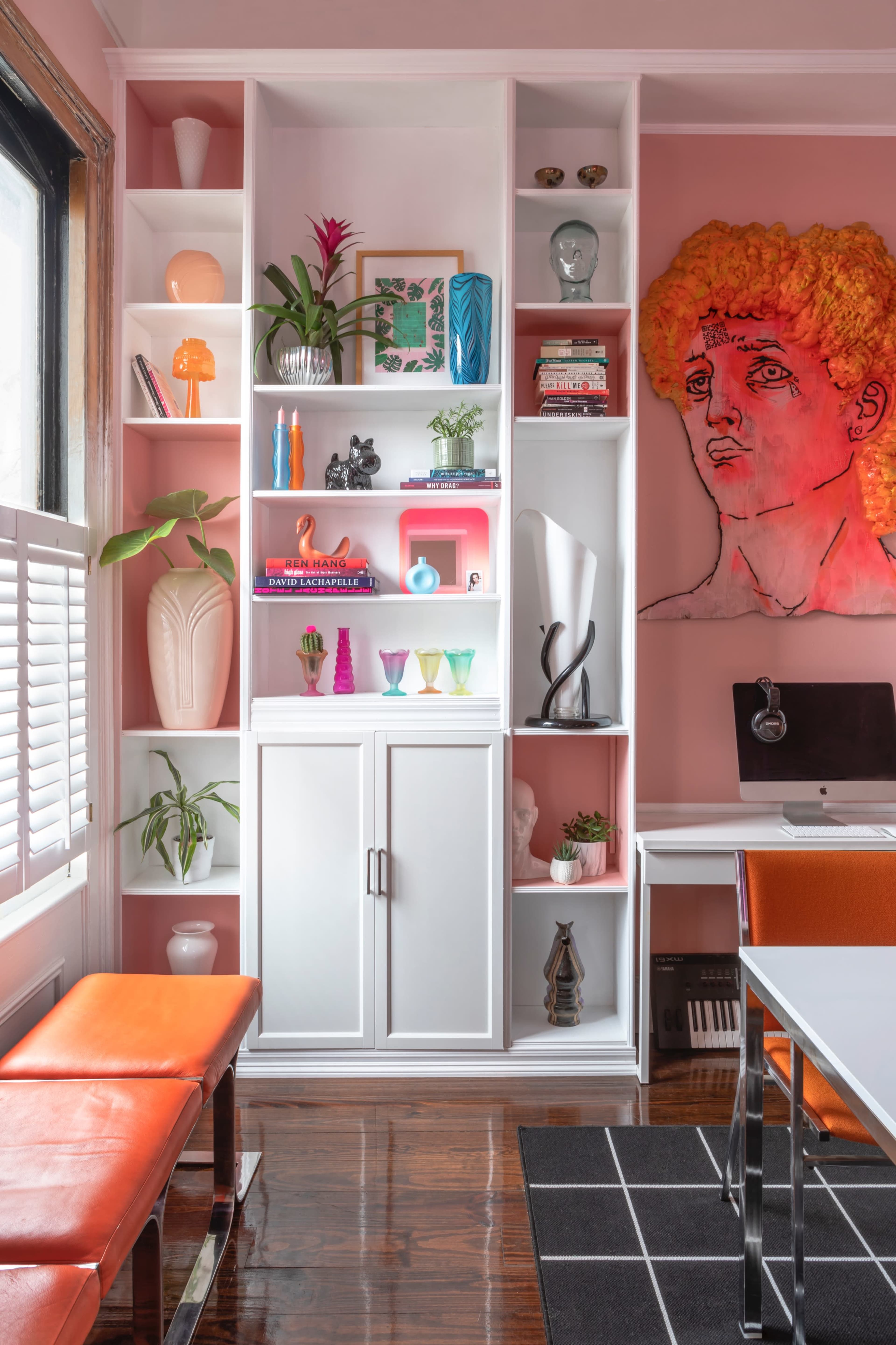 A brightly colored room features a white bookshelf filled with decorative items and a desk with a computer beside orange benches.