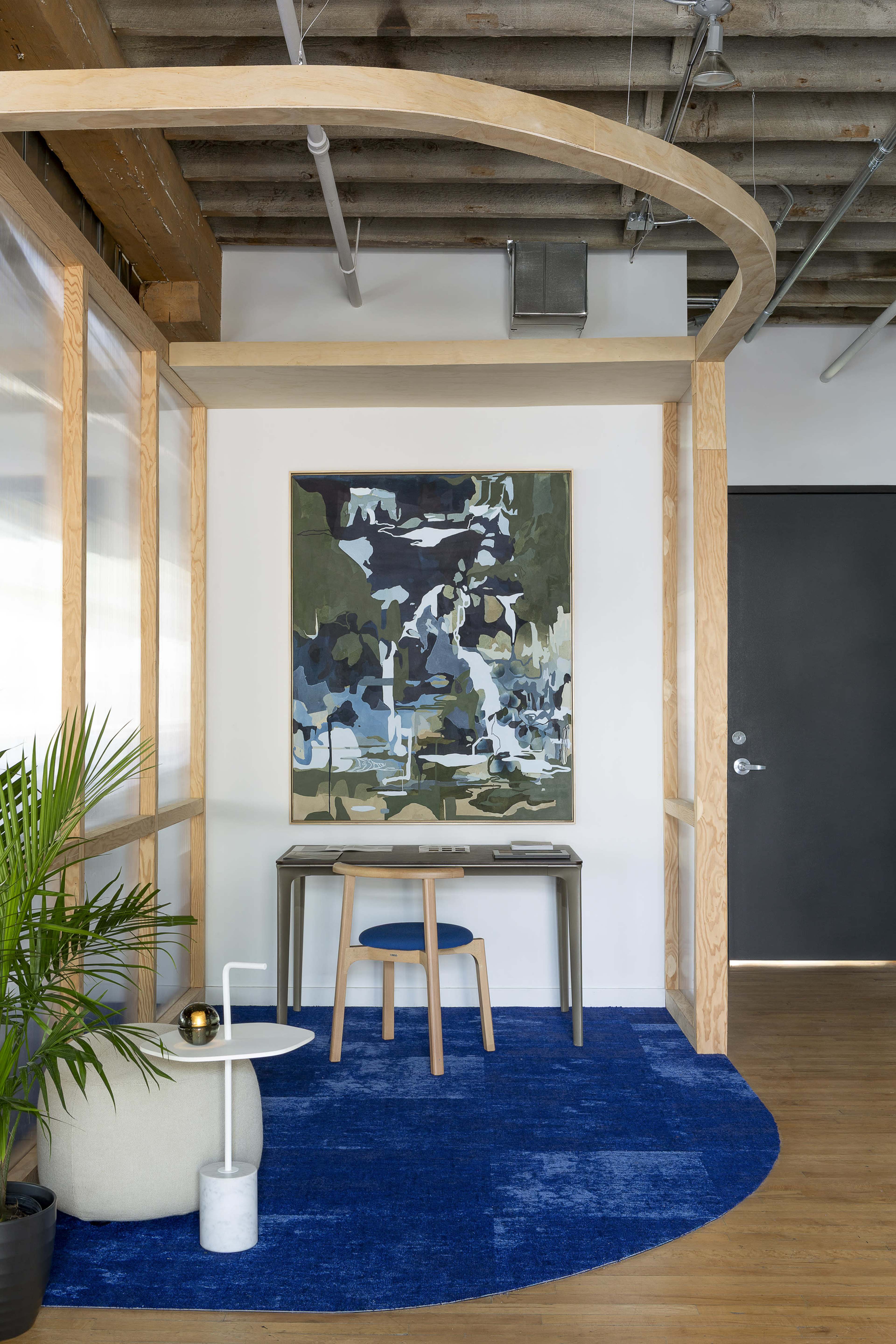 The image features a modern office corner with a wooden desk, a blue area rug, a large abstract painting on the wall, and a potted plant.