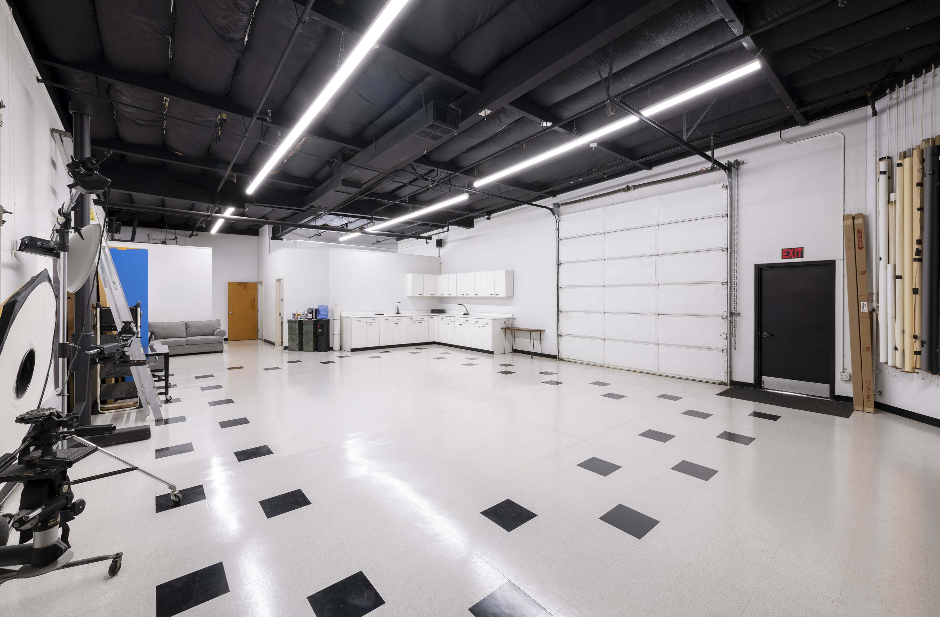 A spacious, empty studio with a black and white tiled floor, white walls, and overhead lighting, featuring cabinets along one wall and a large garage door.