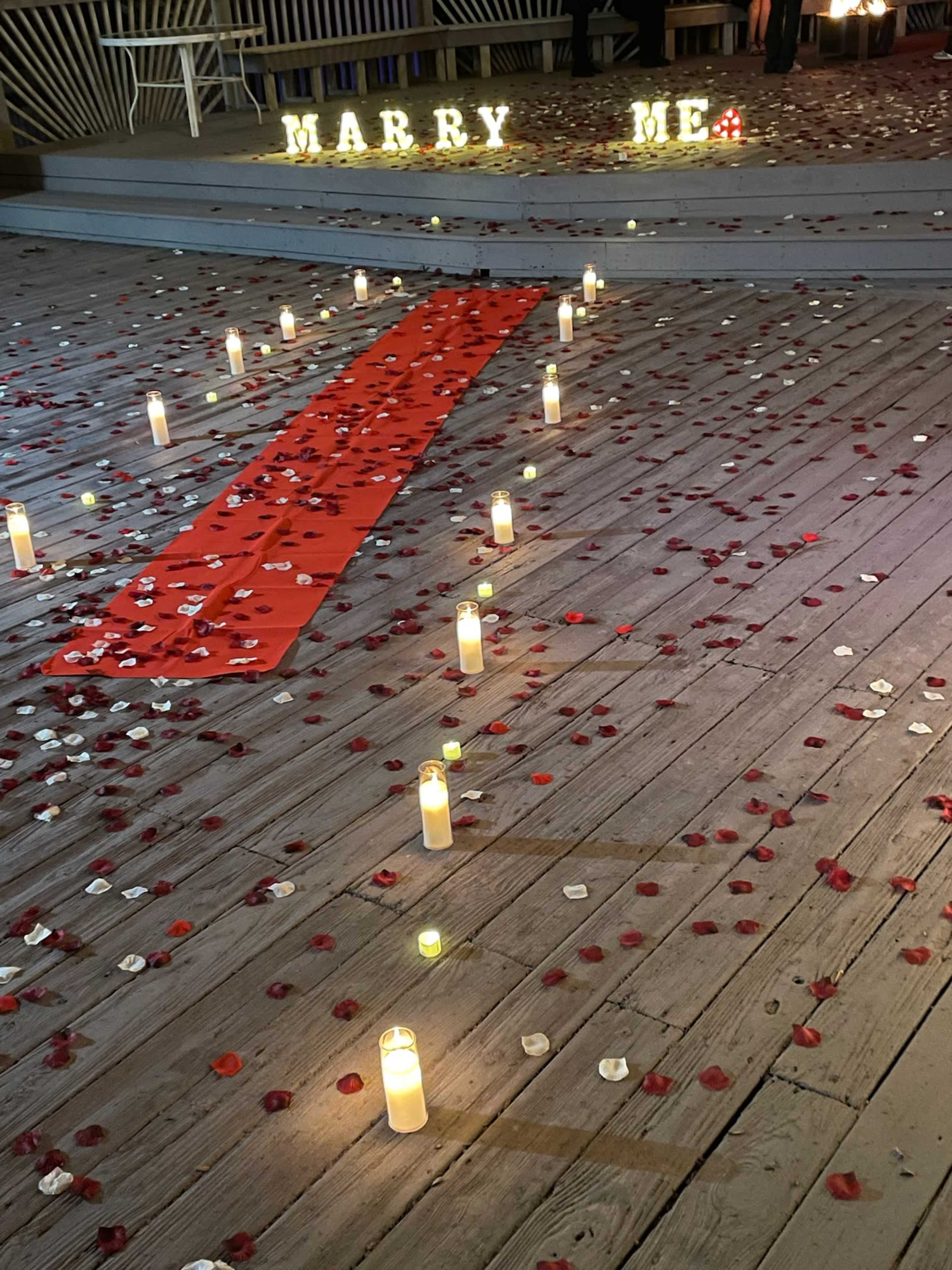 The image shows a romantic proposal setup featuring candles, rose petals, and a red carpet leading up to illuminated letters that spell "MARRY ME."