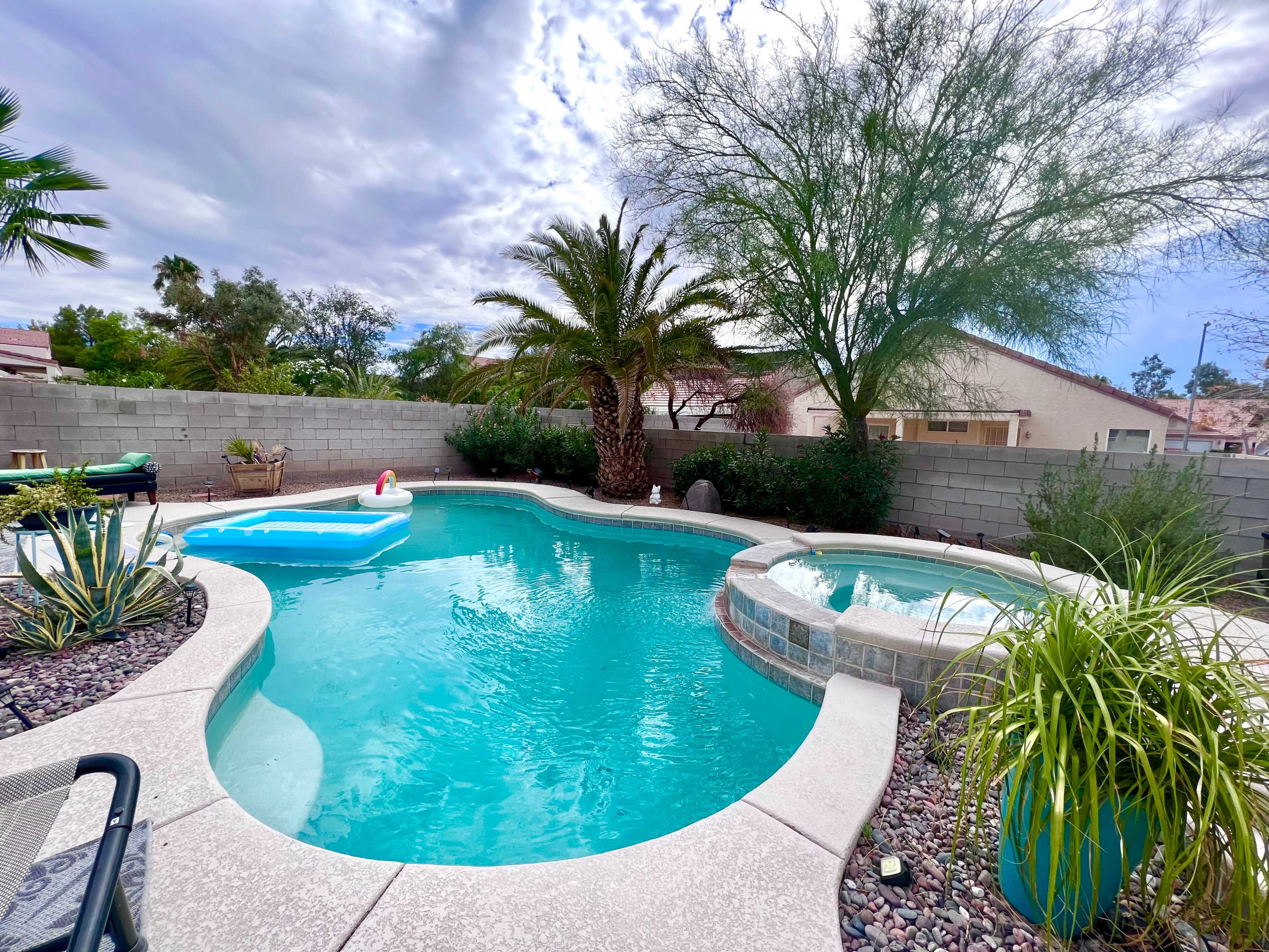 A backyard pool area features a swimming pool with a lounge float, a spa, palm trees, and surrounding landscaping.