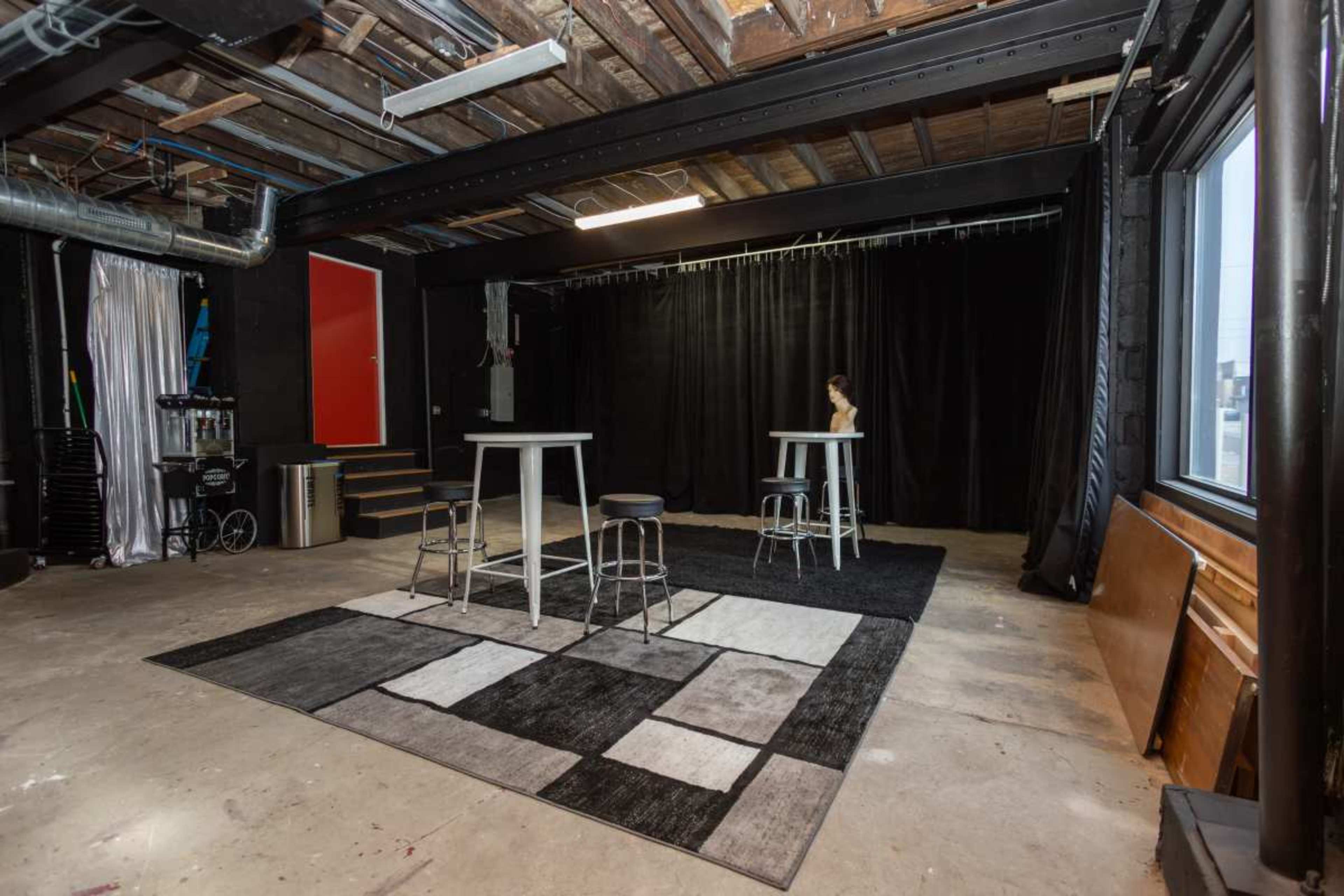 The image shows a spacious studio with black curtains, a concrete floor, two tall white tables, three metal stools, and a large area rug with a geometric pattern.