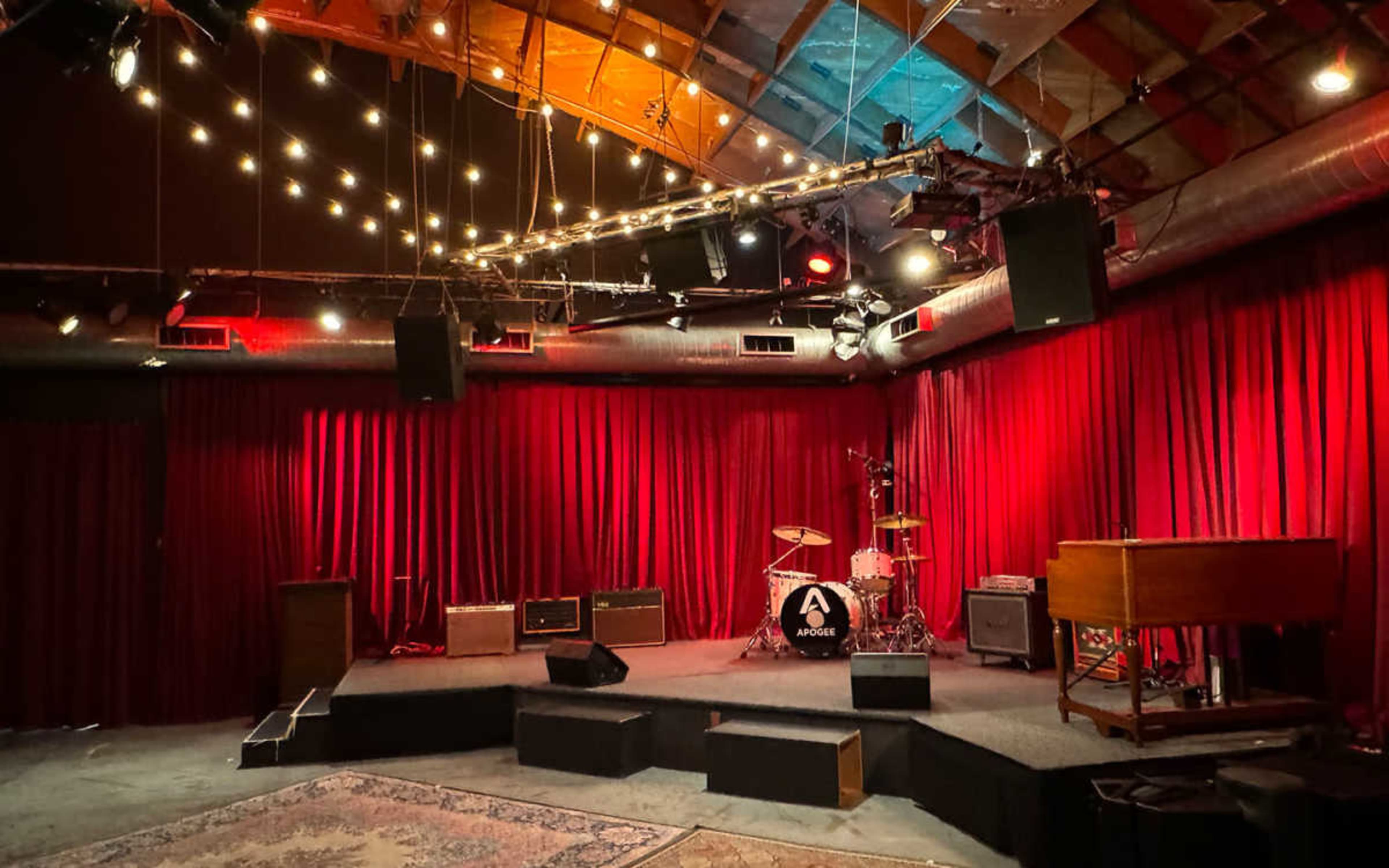 The image depicts a dimly lit music venue with a small stage, red curtains, and various musical instruments, including drums and an organ.