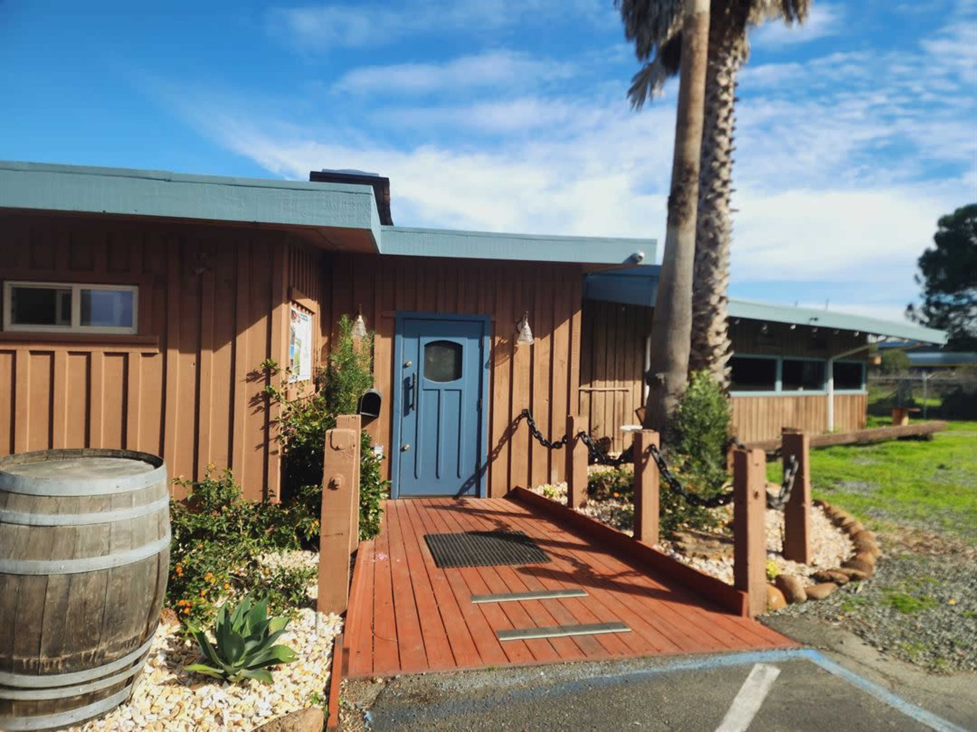 The image shows a wooden building with a blue door and a ramp leading to the entrance, surrounded by landscaping and a palm tree.