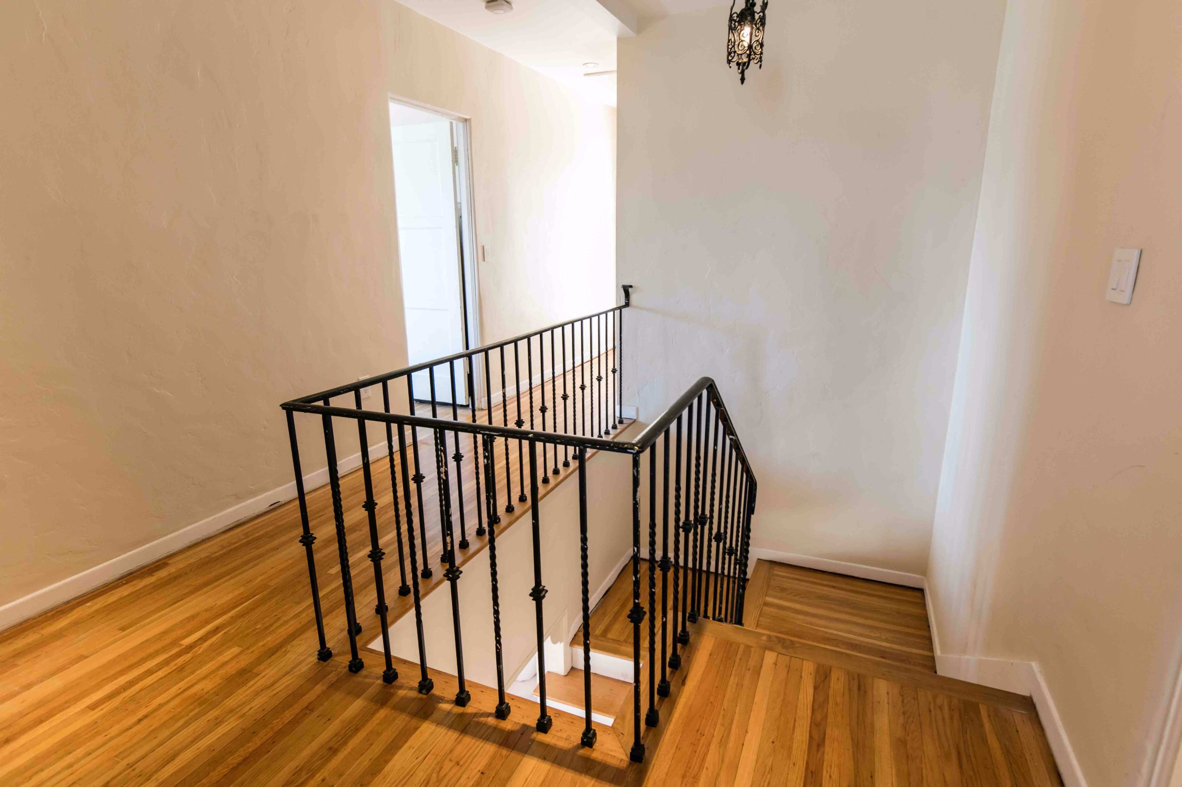 The image shows a wooden staircase with black iron railings leading to an upper level in a well-lit hallway.