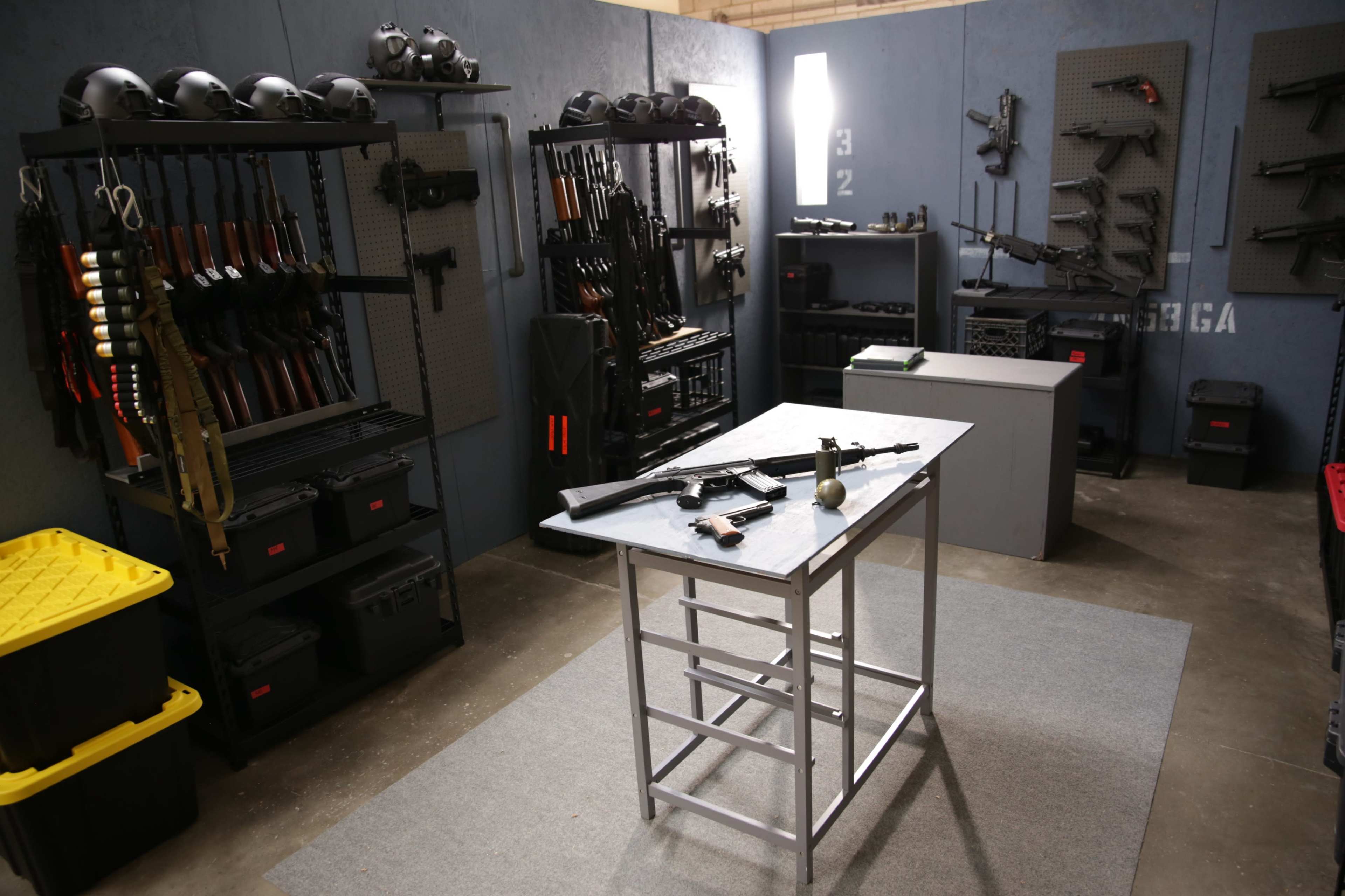 The image shows a room lined with various firearms displayed on racks, with a metal table in the center holding a few weapons and an object, set against a blue wall.