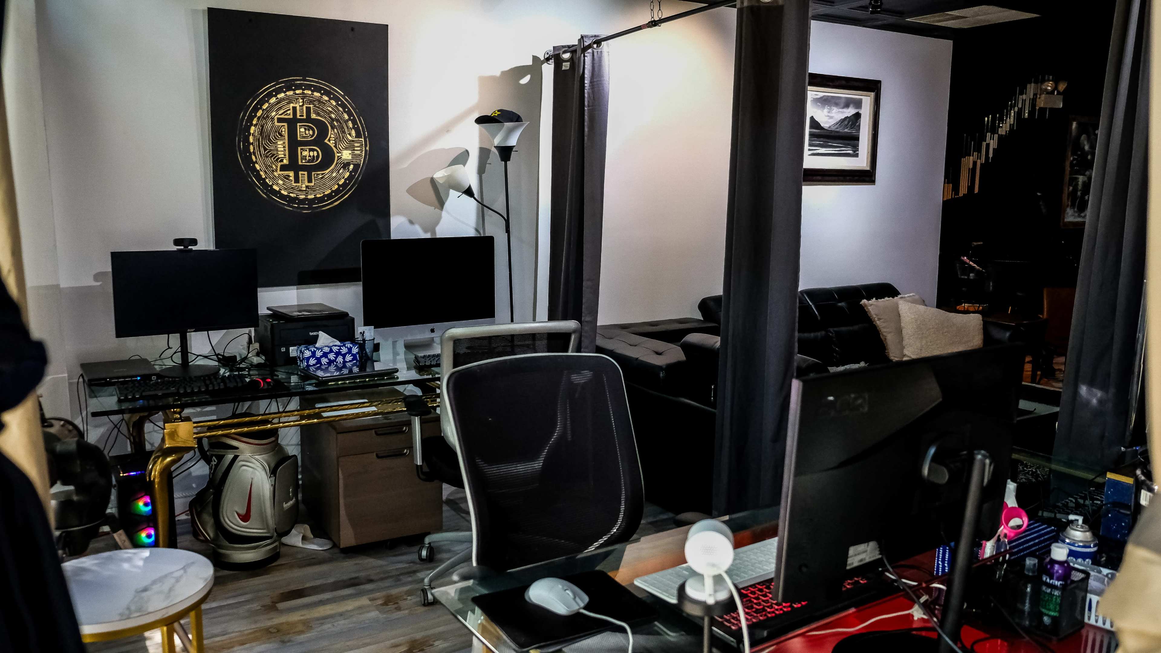 The image shows a modern office space with two computer monitors, a Bitcoin-themed wall decoration, and a black leather couch in the background.