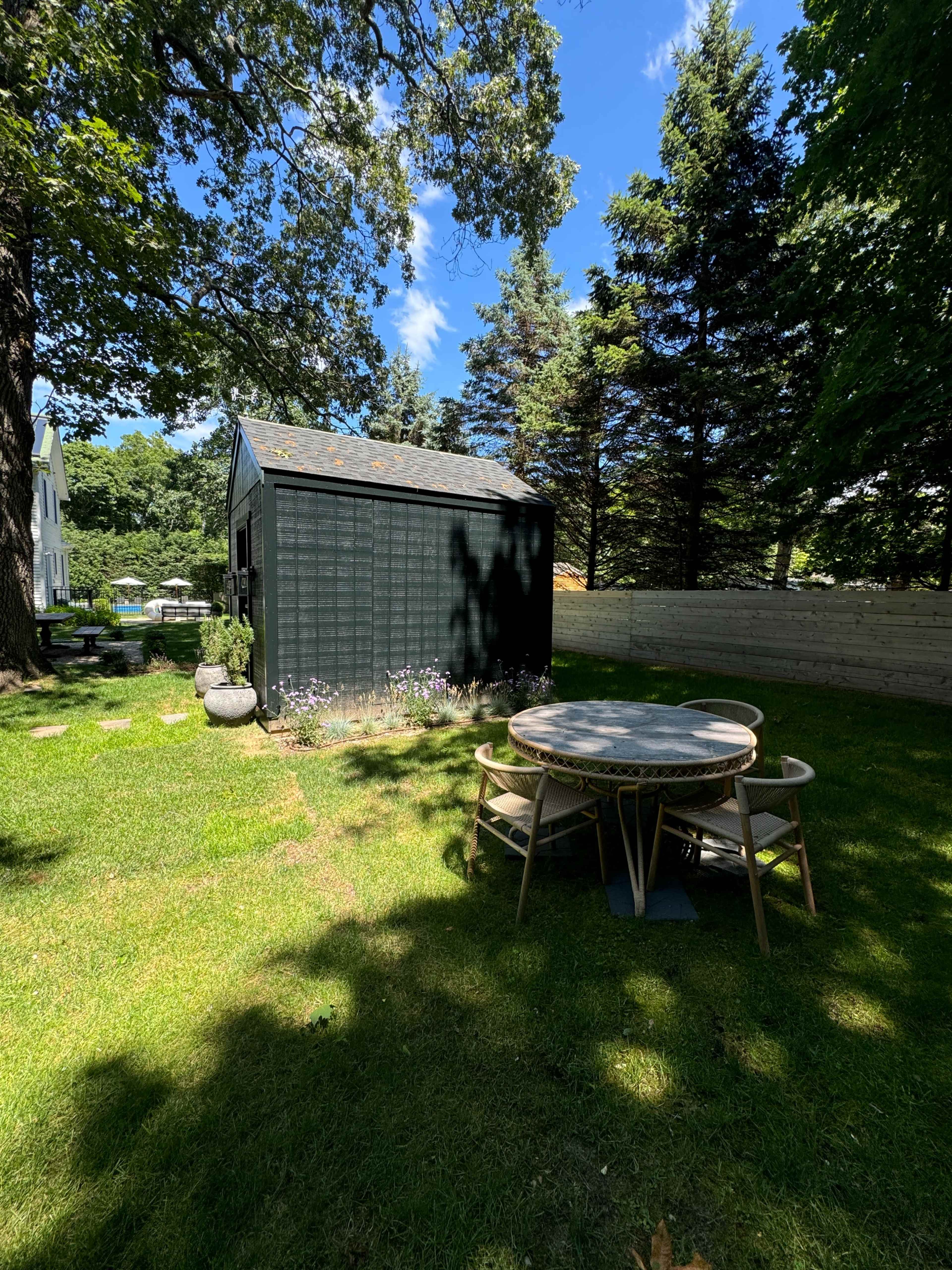 A small black shed stands in a green yard surrounded by trees, with a round table and chairs nearby.