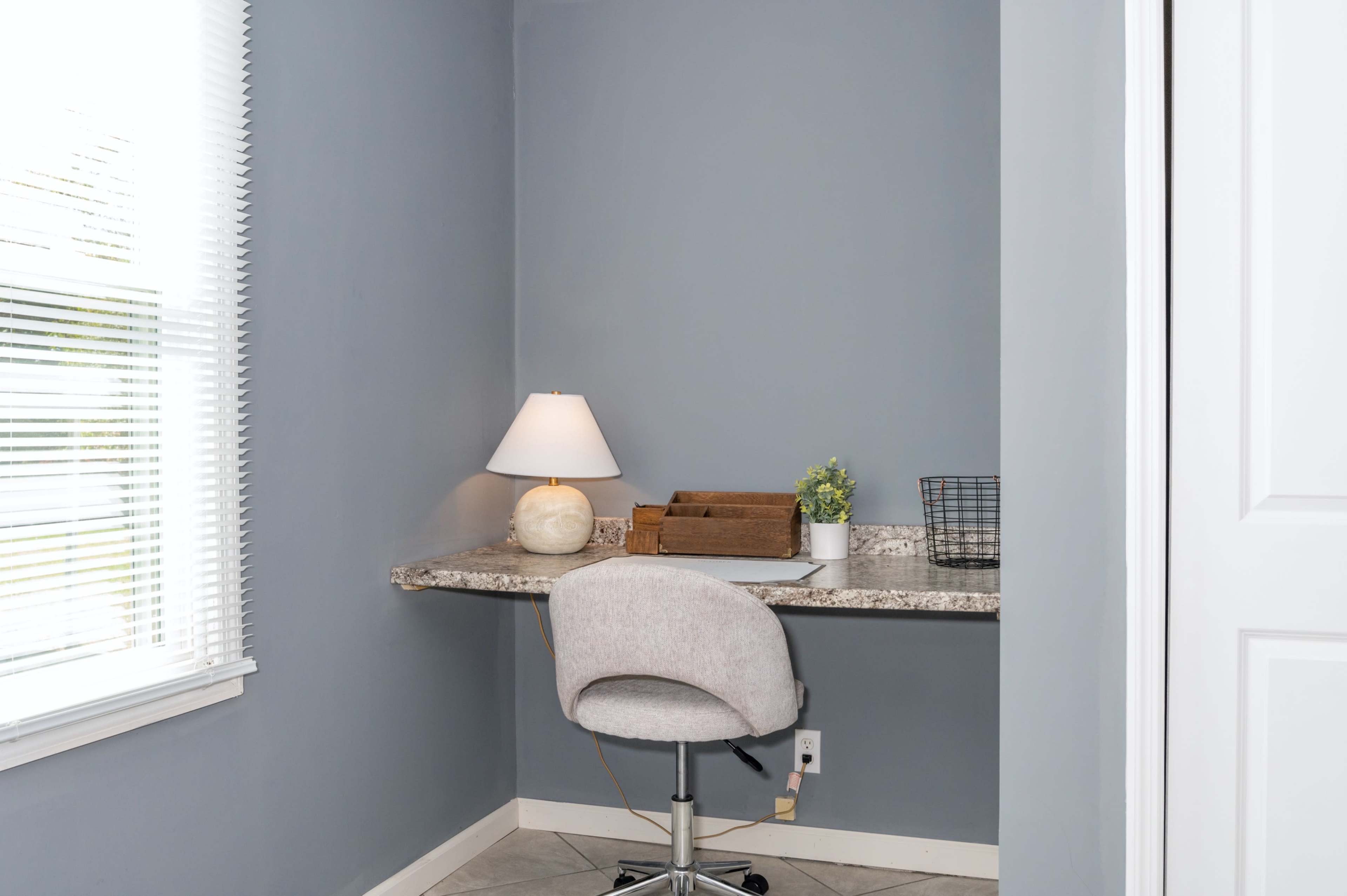 A small home workspace with a gray wall, a desk with a lamp, a wooden organizer, a potted plant, and a mesh basket.