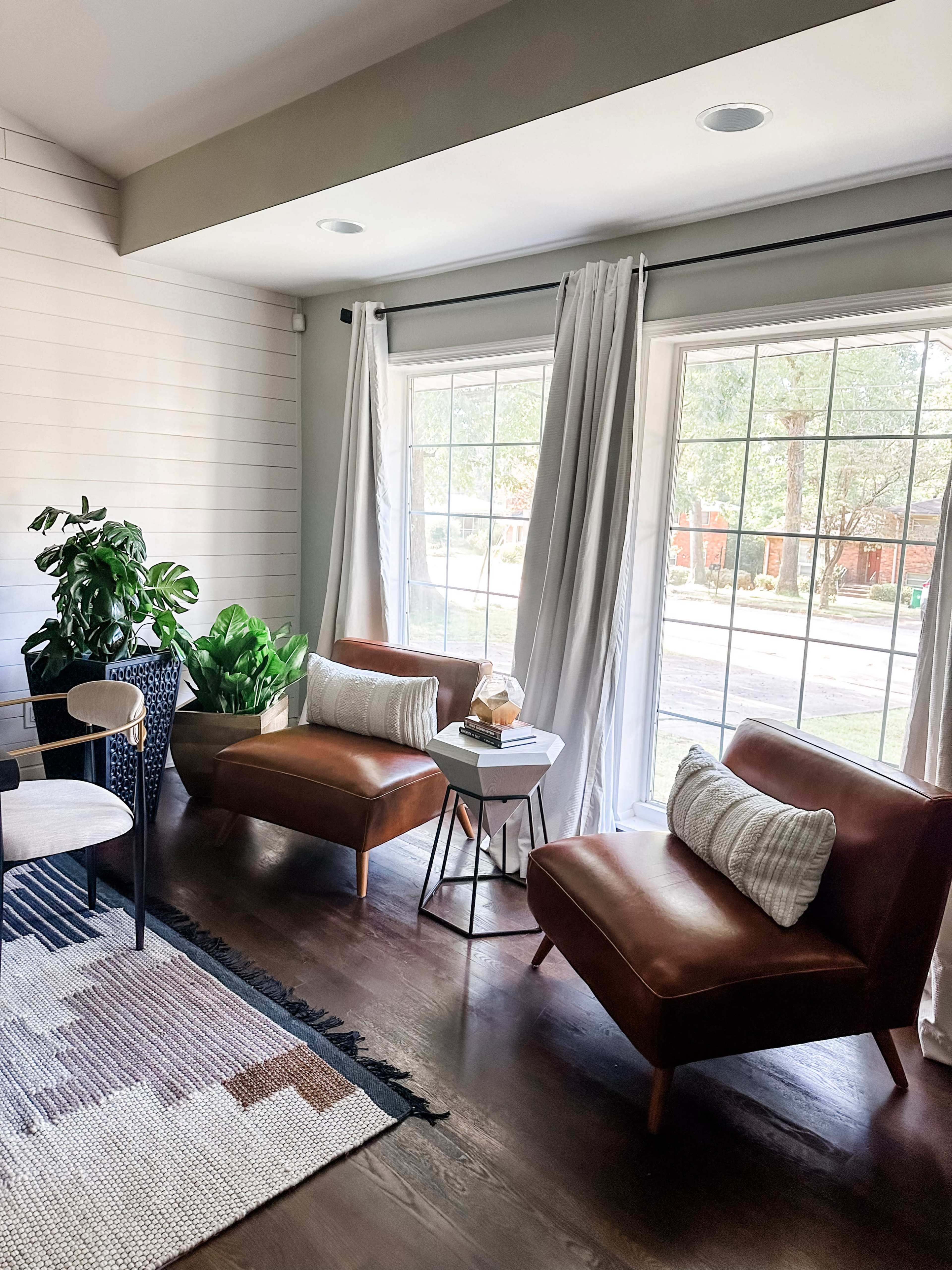 The scene depicts a sunlit living room with two brown armchairs, a small side table between them, and large windows dressed with white curtains.