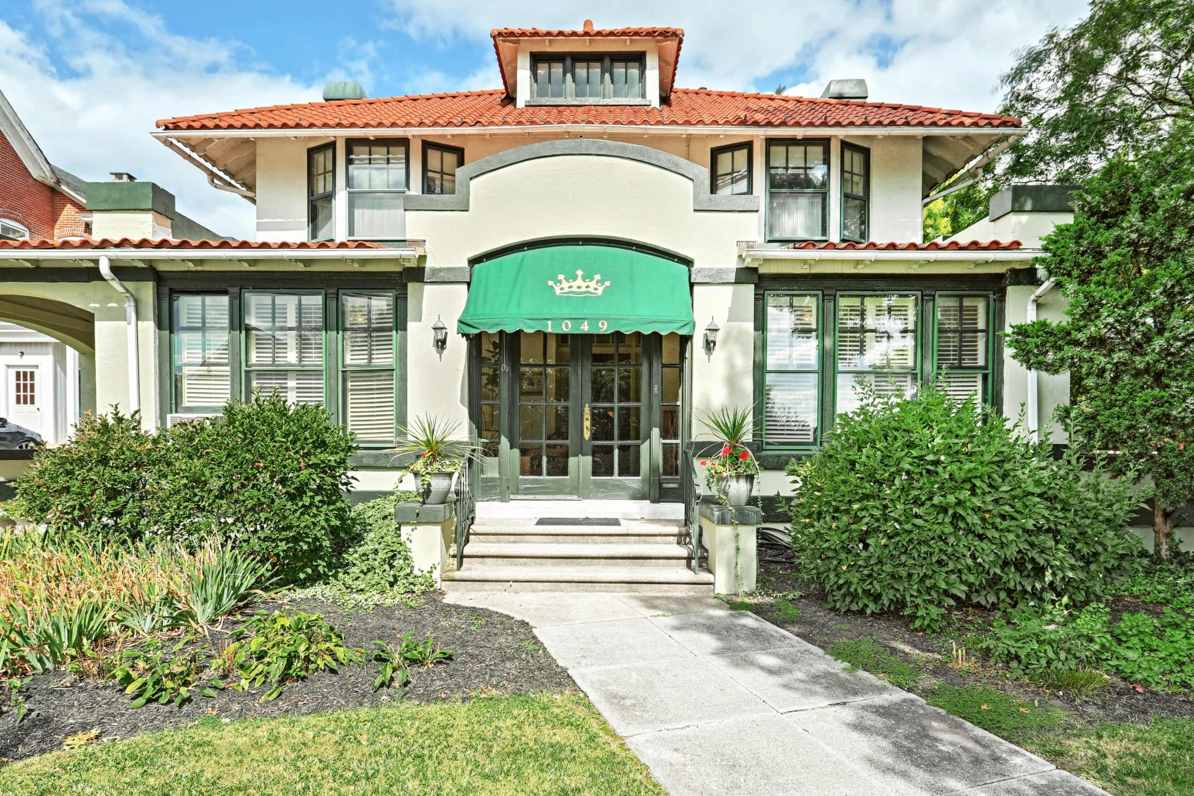 A two-story house with a red tile roof and a green awning, featuring symmetrical windows and well-maintained landscaping, stands prominently at the address 1049.