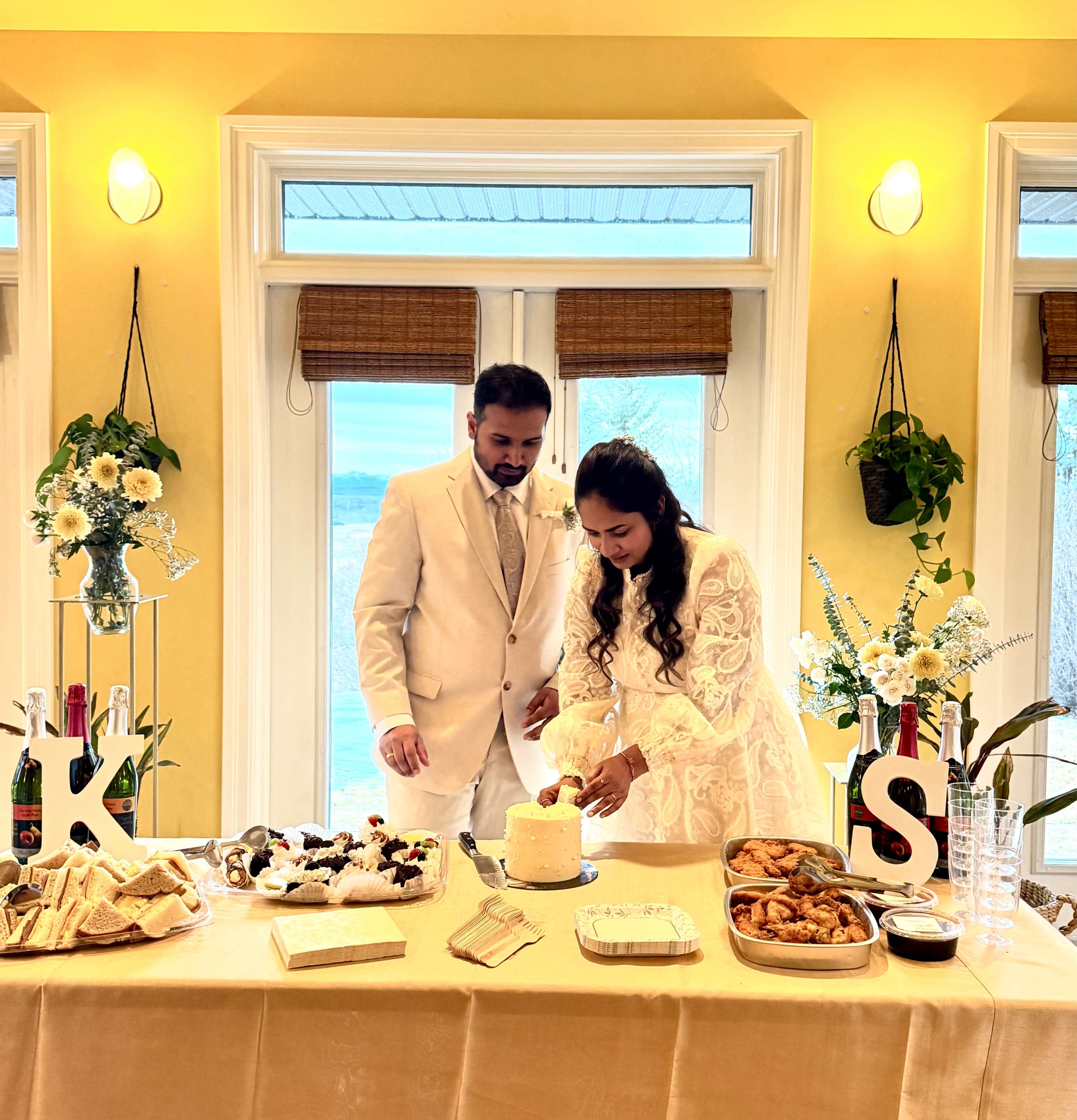 A couple in formal attire cuts a small cake at a decorated table featuring various desserts and snacks.